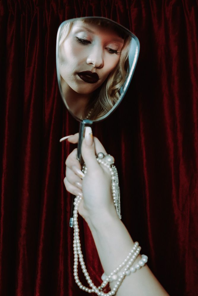 A hand adorned with pearl bracelet holds a mirror reflecting a blonde woman with dark lipstick against red velvet.