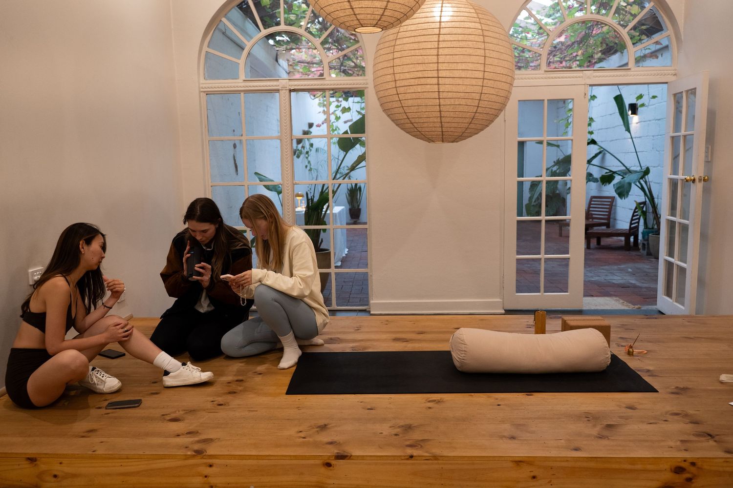 Three women sit on a yoga mat in a bright studio with French doors, large windows, and a paper lantern overhead.