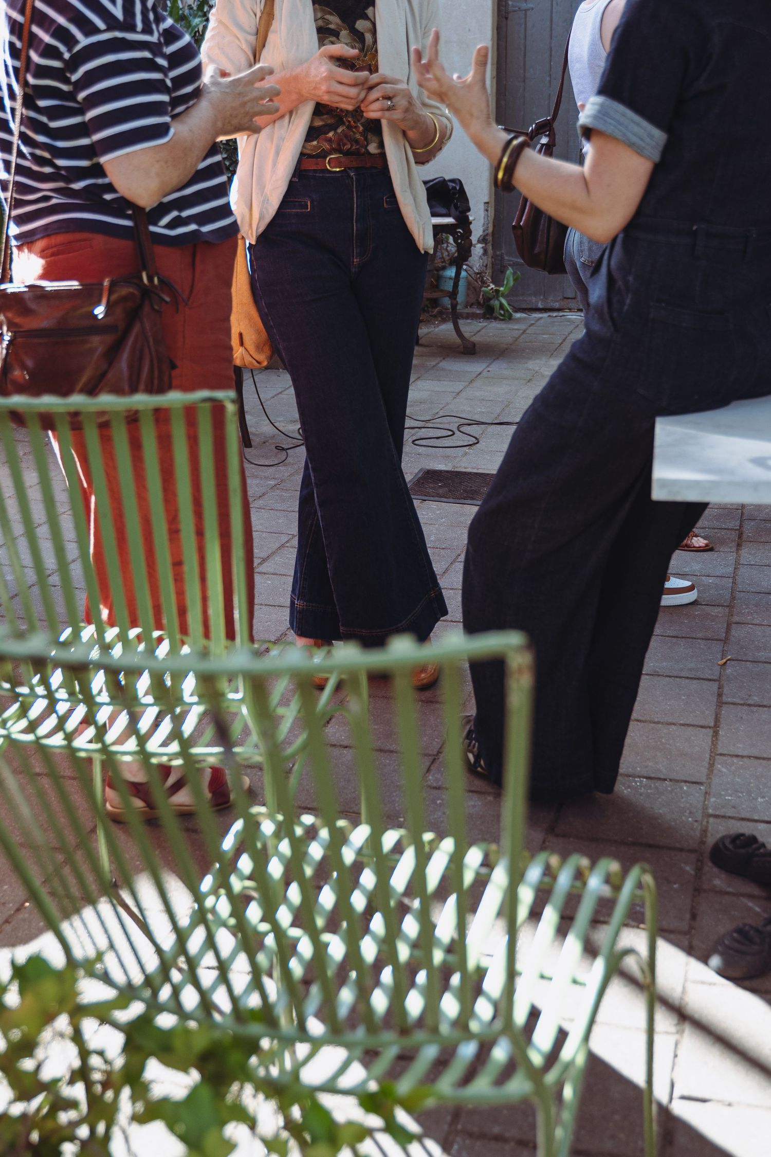 People socializing outdoors on a brick patio with green metal chairs, wearing casual flared jeans and overalls.