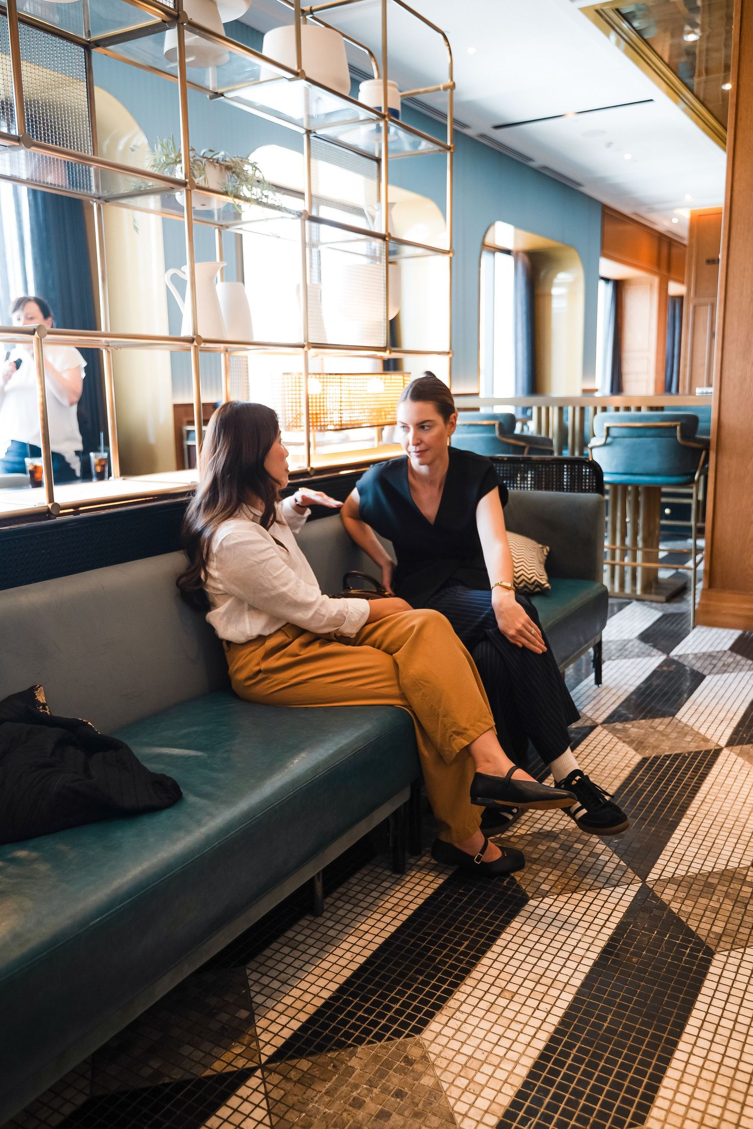 Two women sitting on a teal sofa in a stylish restaurant lounge, engaged in conversation.