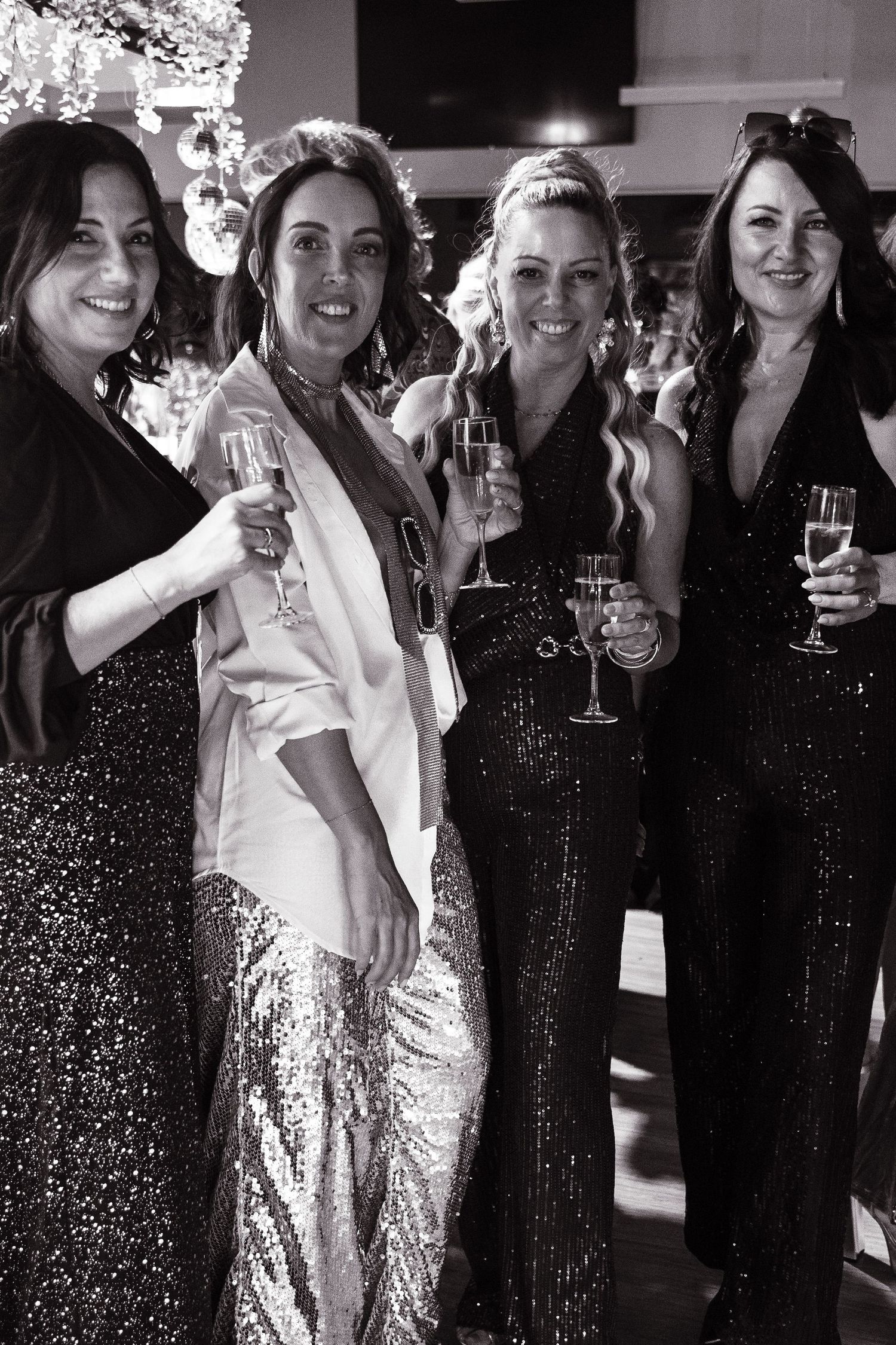 Four women in sequin gowns smiling and holding champagne glasses at a formal event, black and white photo.