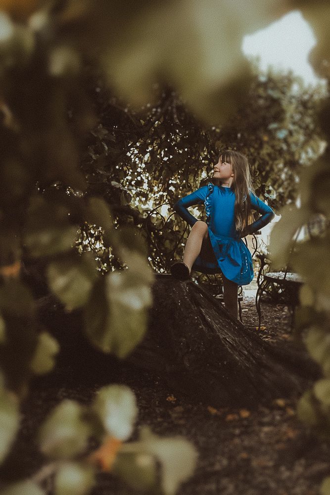 Woman in blue dress poses gracefully among ancient trees in a moody, atmospheric forest setting.