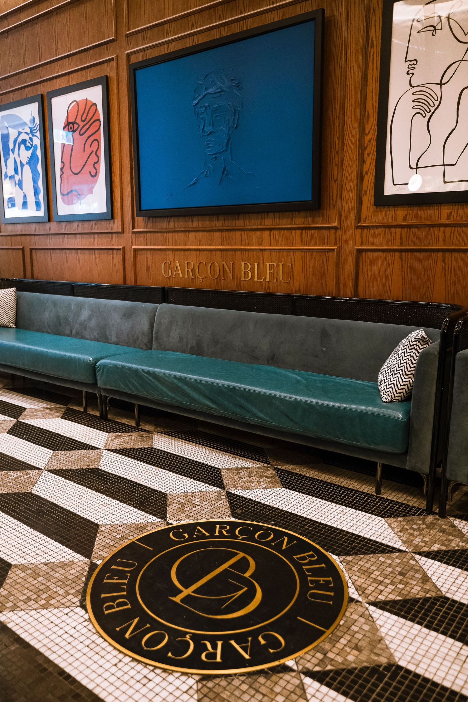Garcon Bleu restaurant interior with teal banquette seating, checkered tile floor logo, wood paneling, and modern art prints.