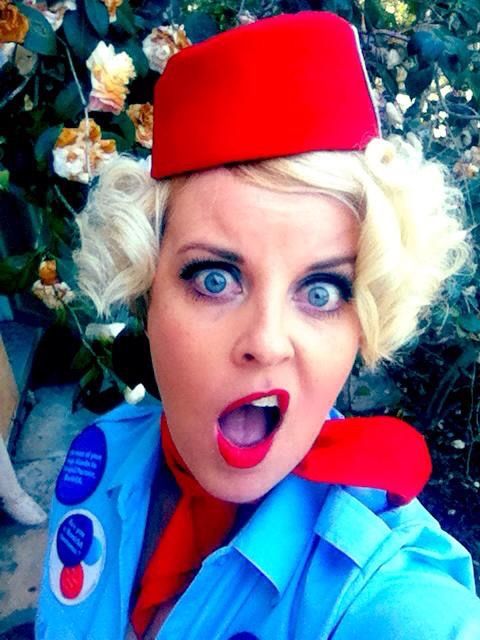 Woman in retro flight attendant costume with red hat, blue uniform, and dramatic makeup poses with surprised expression.