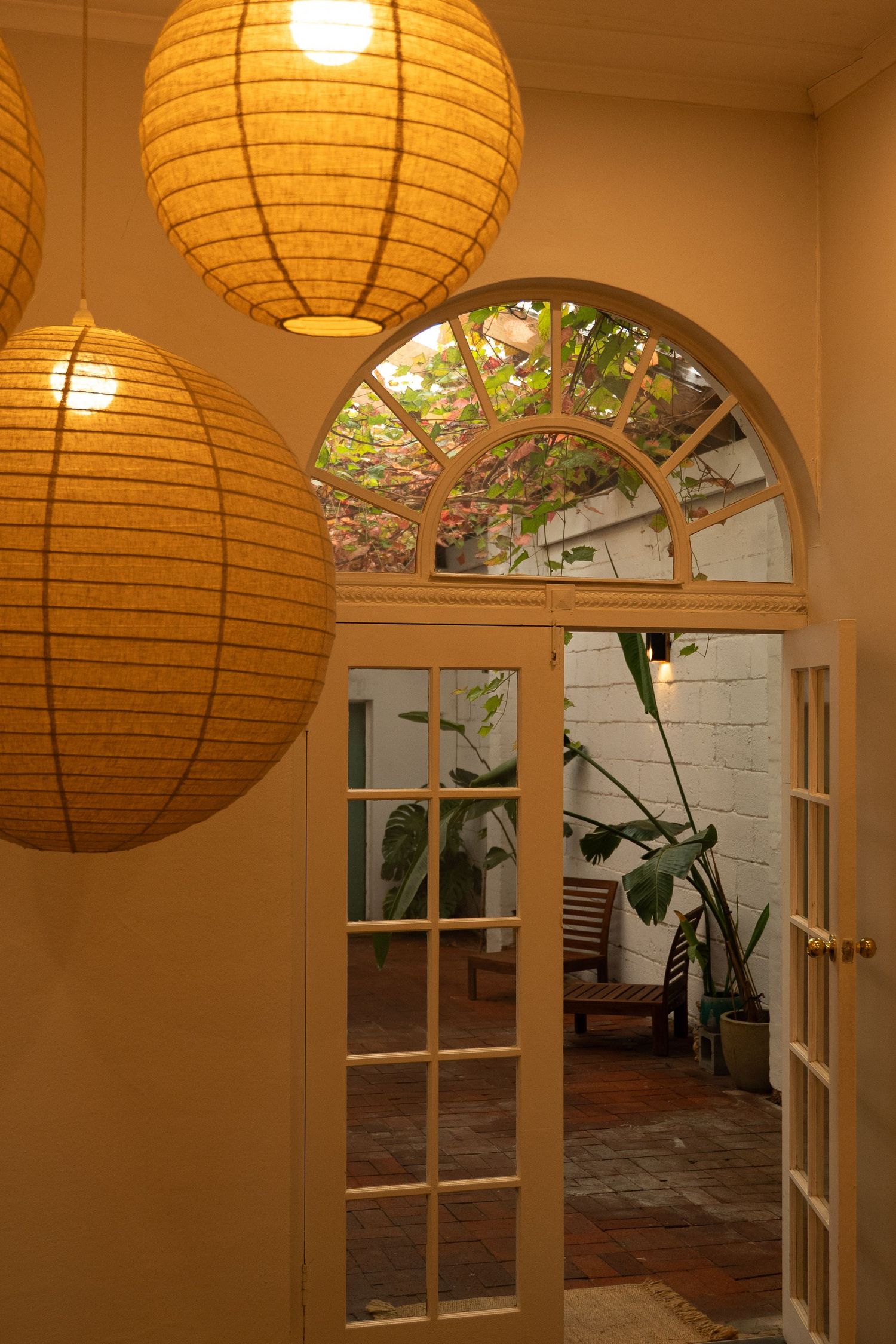 Warm hallway with paper lantern lights leading to French doors opening to a lush courtyard garden.
