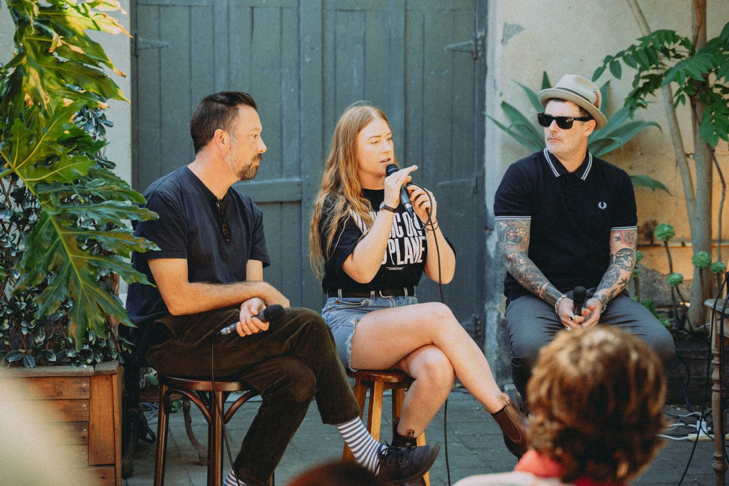 Three people sit outdoors at a panel discussion, with a tattooed woman speaking into a microphone between two men.