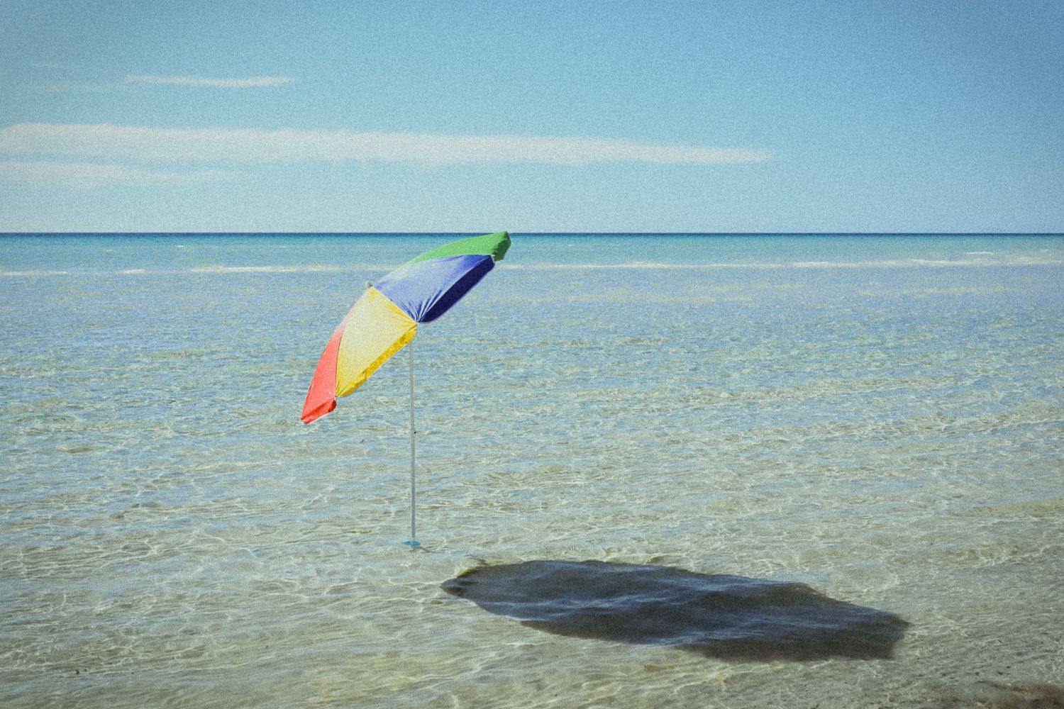 Colorful beach umbrella standing in shallow crystal clear ocean water under a bright blue sky.