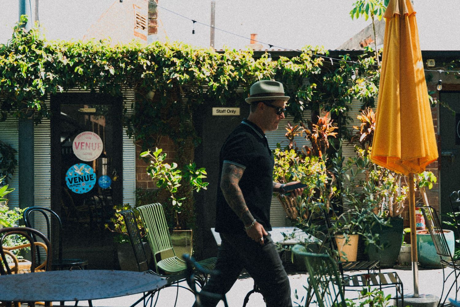 Man in black outfit walking past a lush green cafe patio with outdoor seating and orange umbrella.