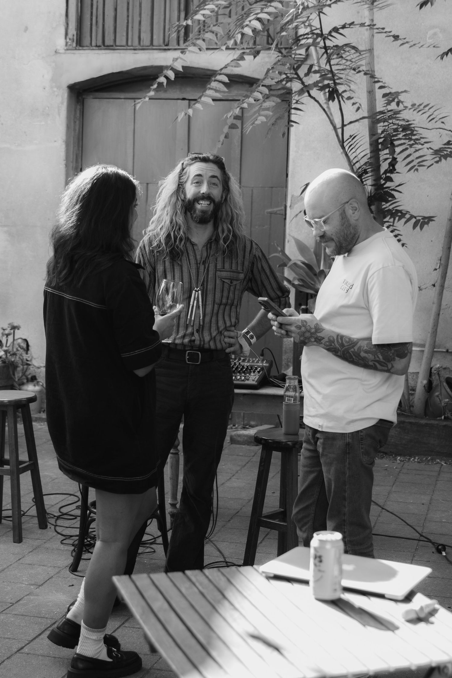 Three people socializing outdoors in black and white photo, two men with long hair and tattooed man checking phone.