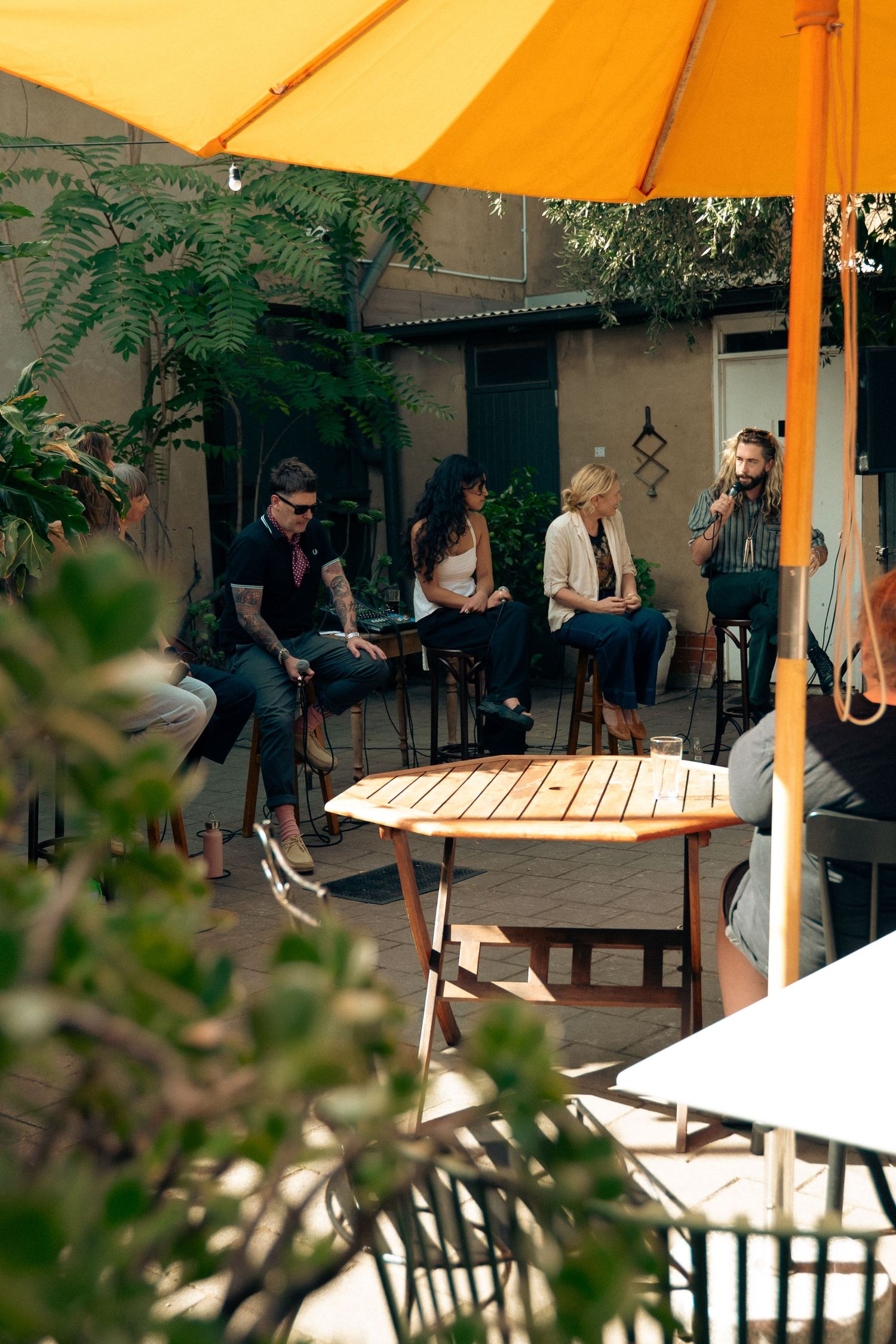 Group of people socializing at an outdoor cafe patio under yellow umbrellas surrounded by lush greenery.