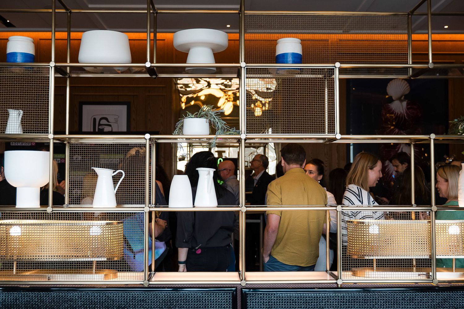Modern shelving display with white ceramic vases and glowing lamps at a stylish indoor event with guests mingling.