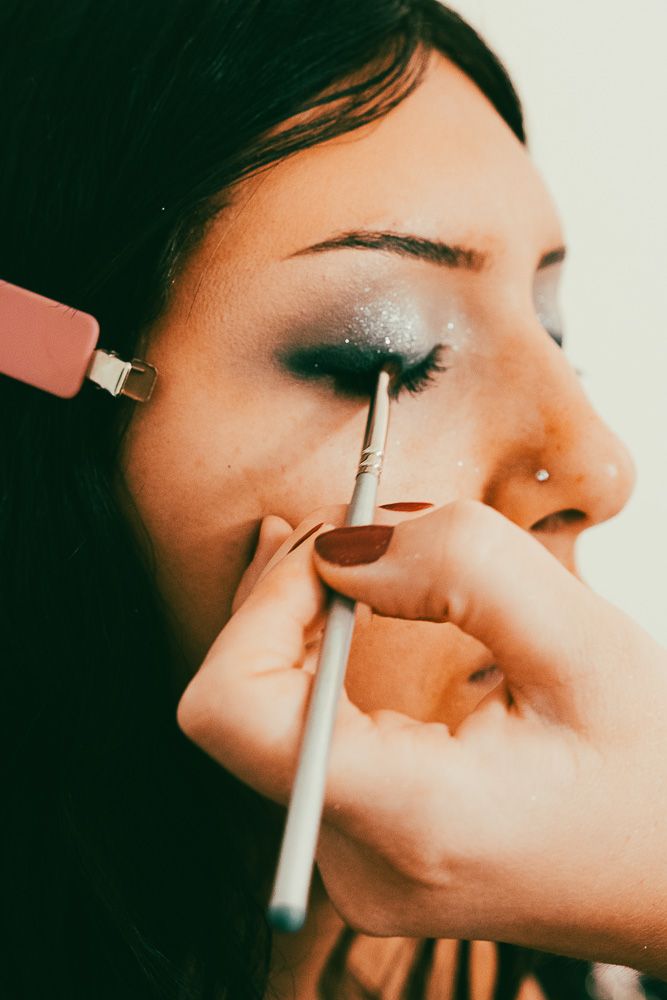 A makeup artist applies glittery smoky eye shadow with a brush on a dark-haired woman with a nose piercing.