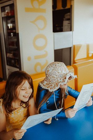 Two young girls sitting at a blue table in a colorful restaurant, smiling and reading menus together.