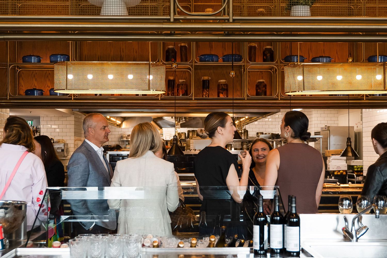 Guests mingle at a stylish restaurant bar with wine bottles, glassware, and an open kitchen in the background.