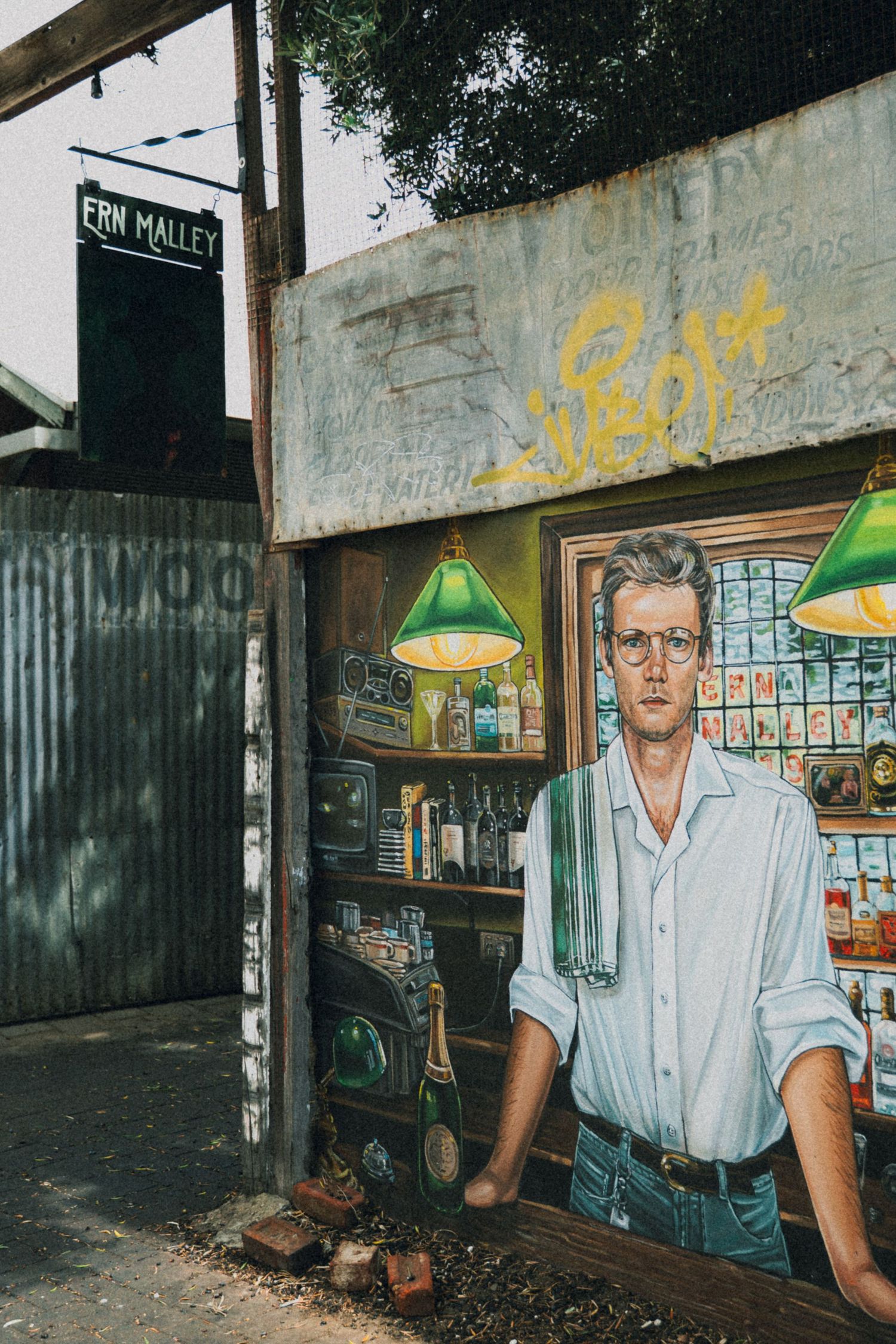 Street mural of a man behind a bar counter with green lamps, near an Ern Malley sign in an alley.
