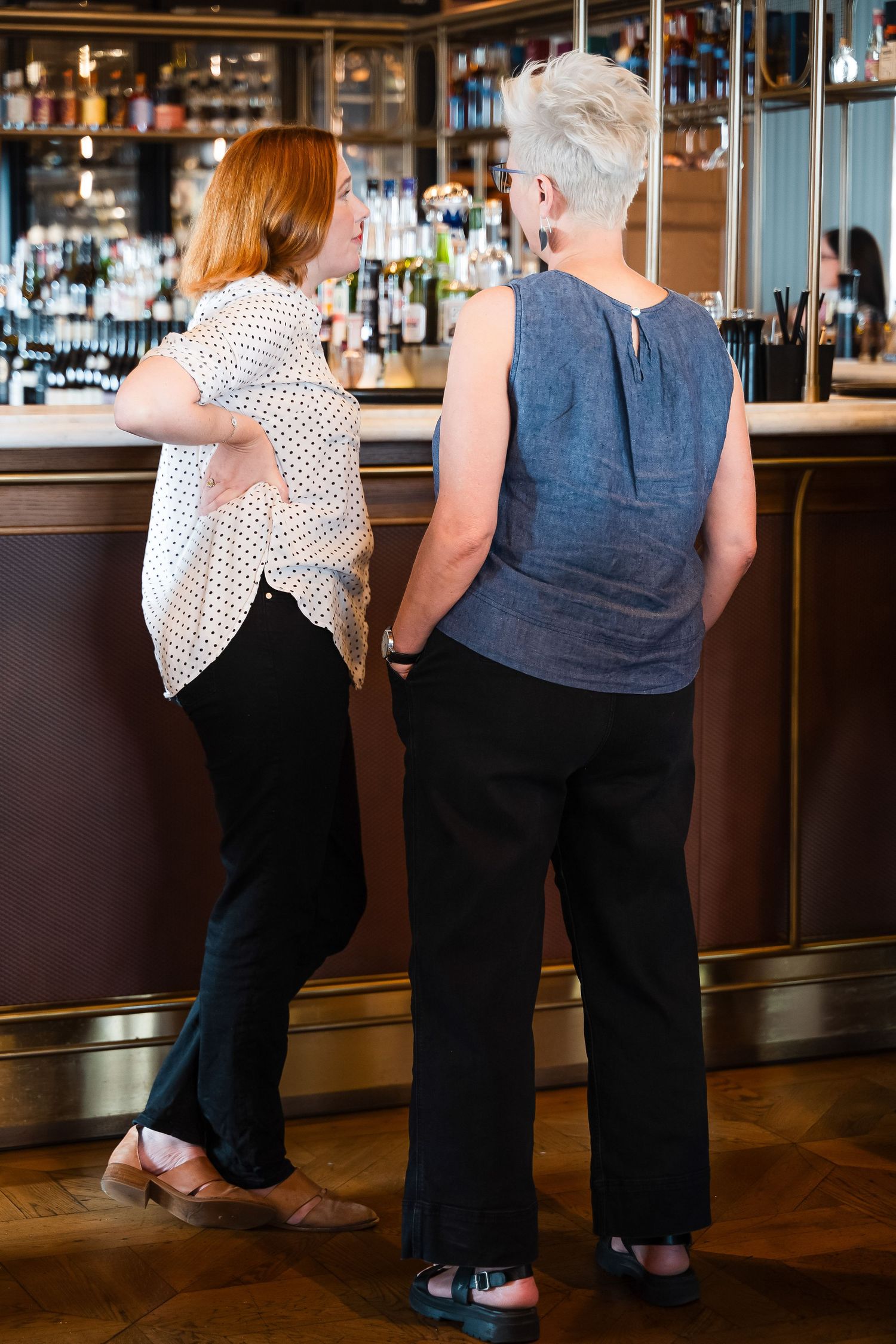 Two women standing at a bar counter, one with red hair in a polka dot blouse, the other with short gray hair in a blue top.