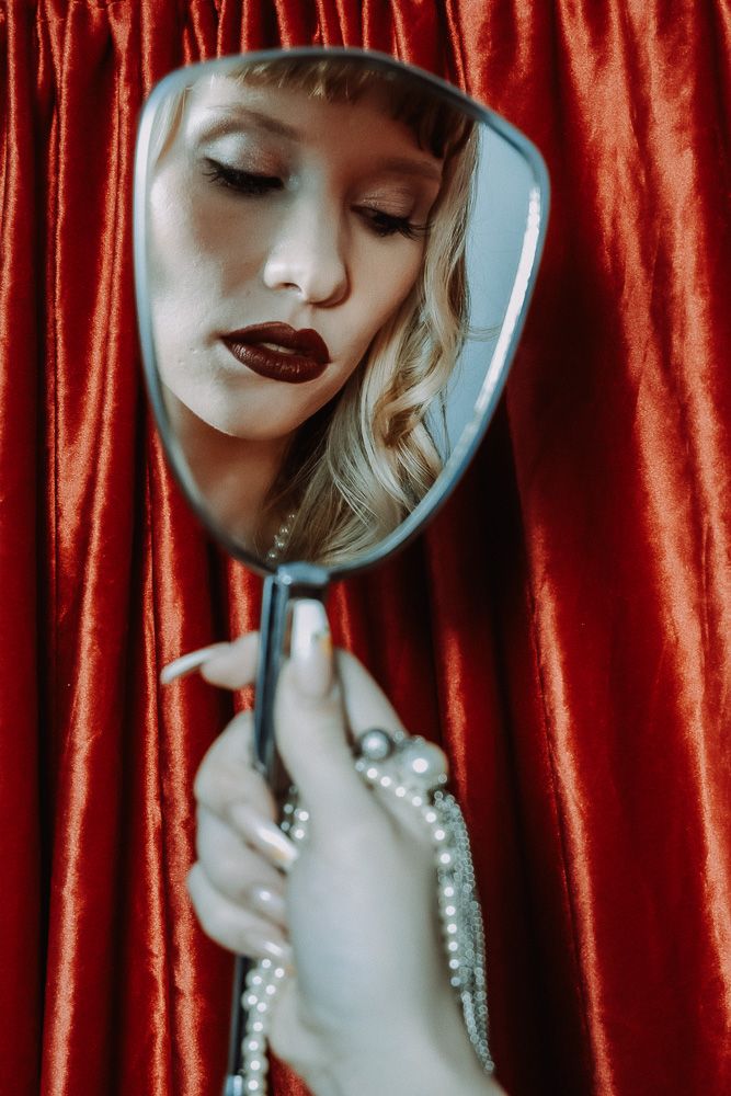 A blonde woman with dark lips gazes into a handheld mirror against a red velvet backdrop.