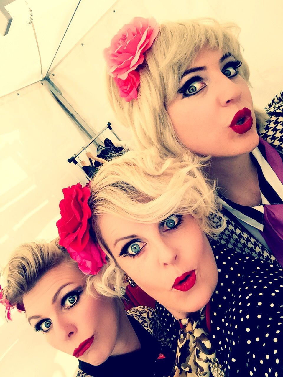 Three women with vintage pin-up style, blonde hair, red lips, and floral accessories pose together for a selfie.