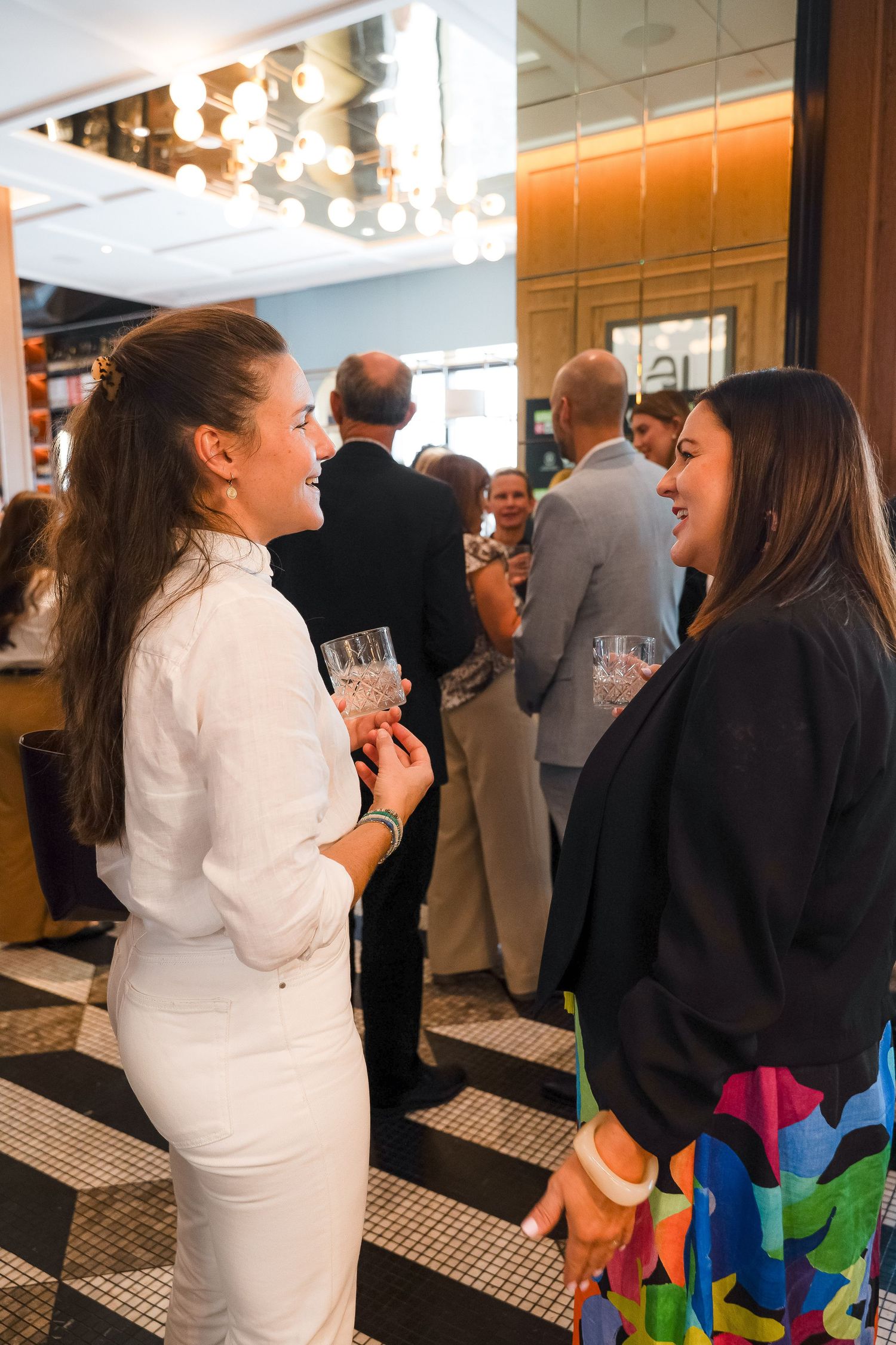 Two women smile and chat at an elegant indoor cocktail party, one in white holding a drink.