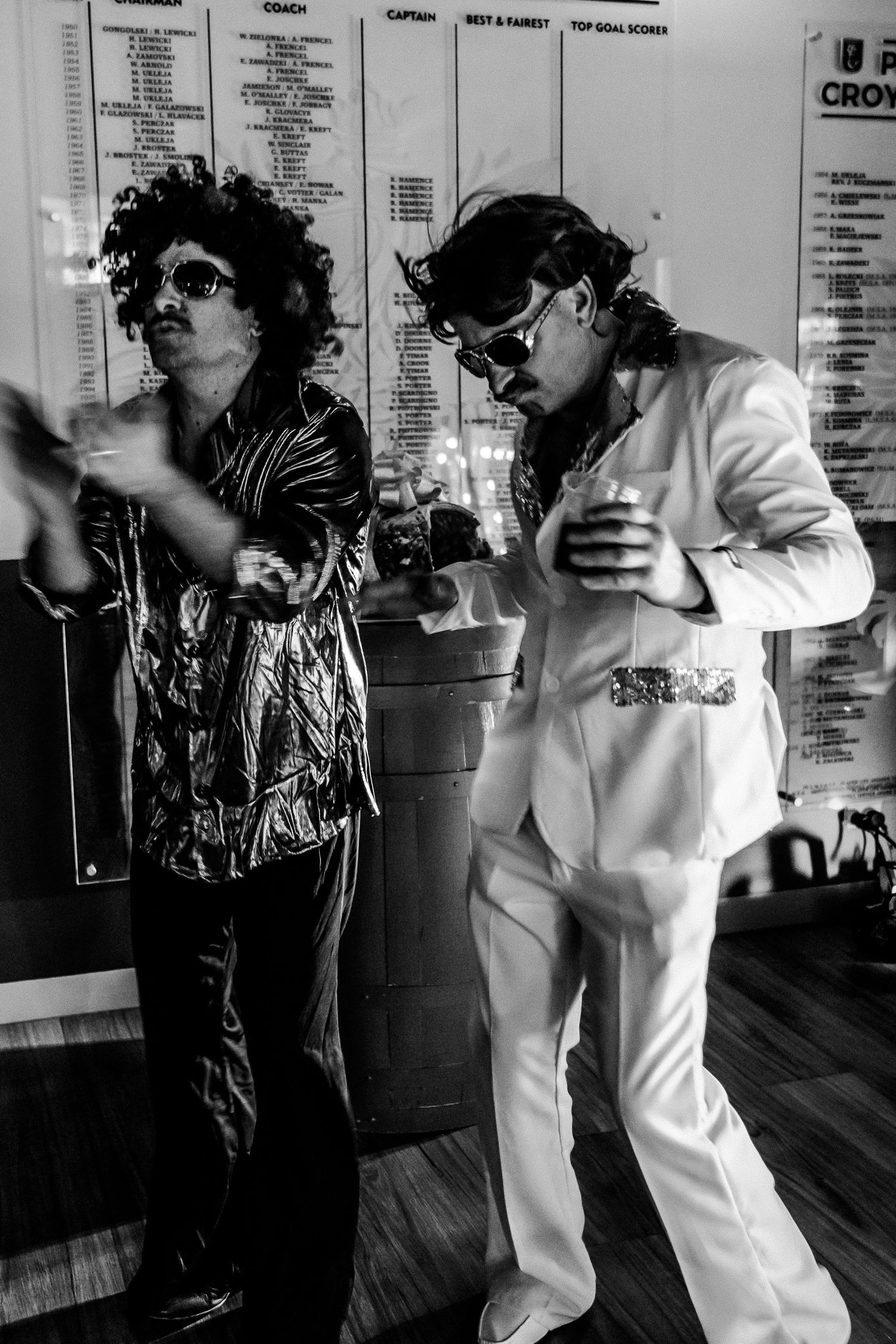 Two men in 70s disco costumes pose at a party, one in gold jacket, one in white suit, both wearing wigs.
