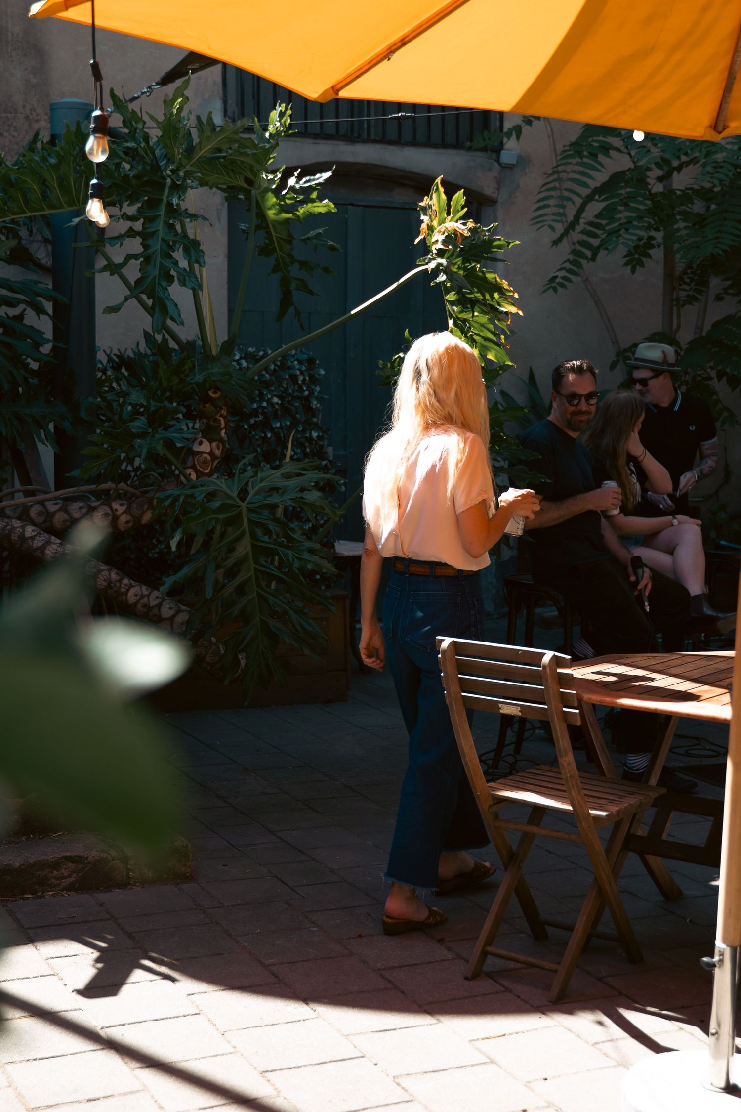Woman in pink top and jeans stands in sunny outdoor cafe patio with yellow umbrella and tropical plants.