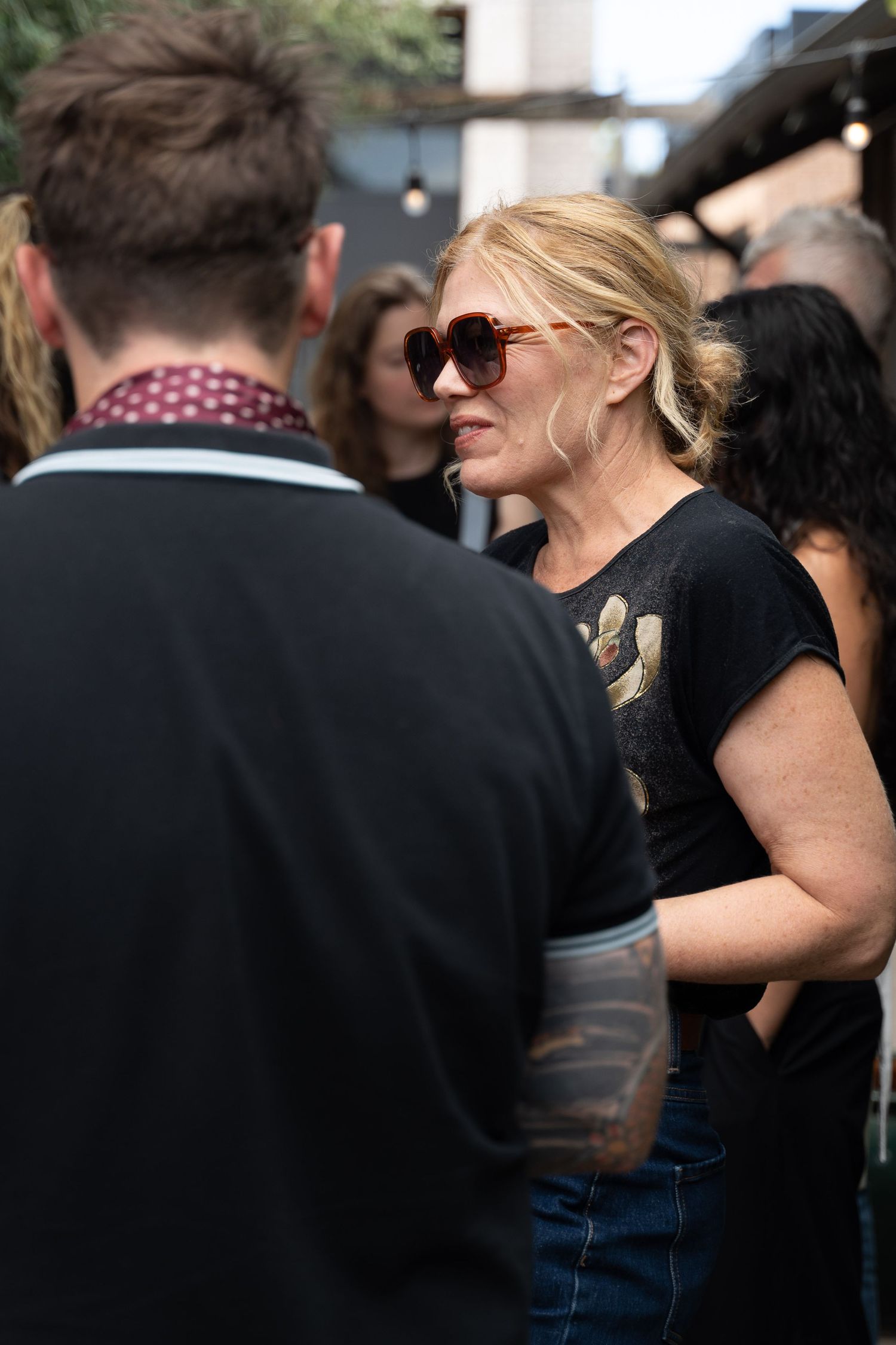 Blonde woman in red sunglasses and black t-shirt socializing at an outdoor event with crowd in background.