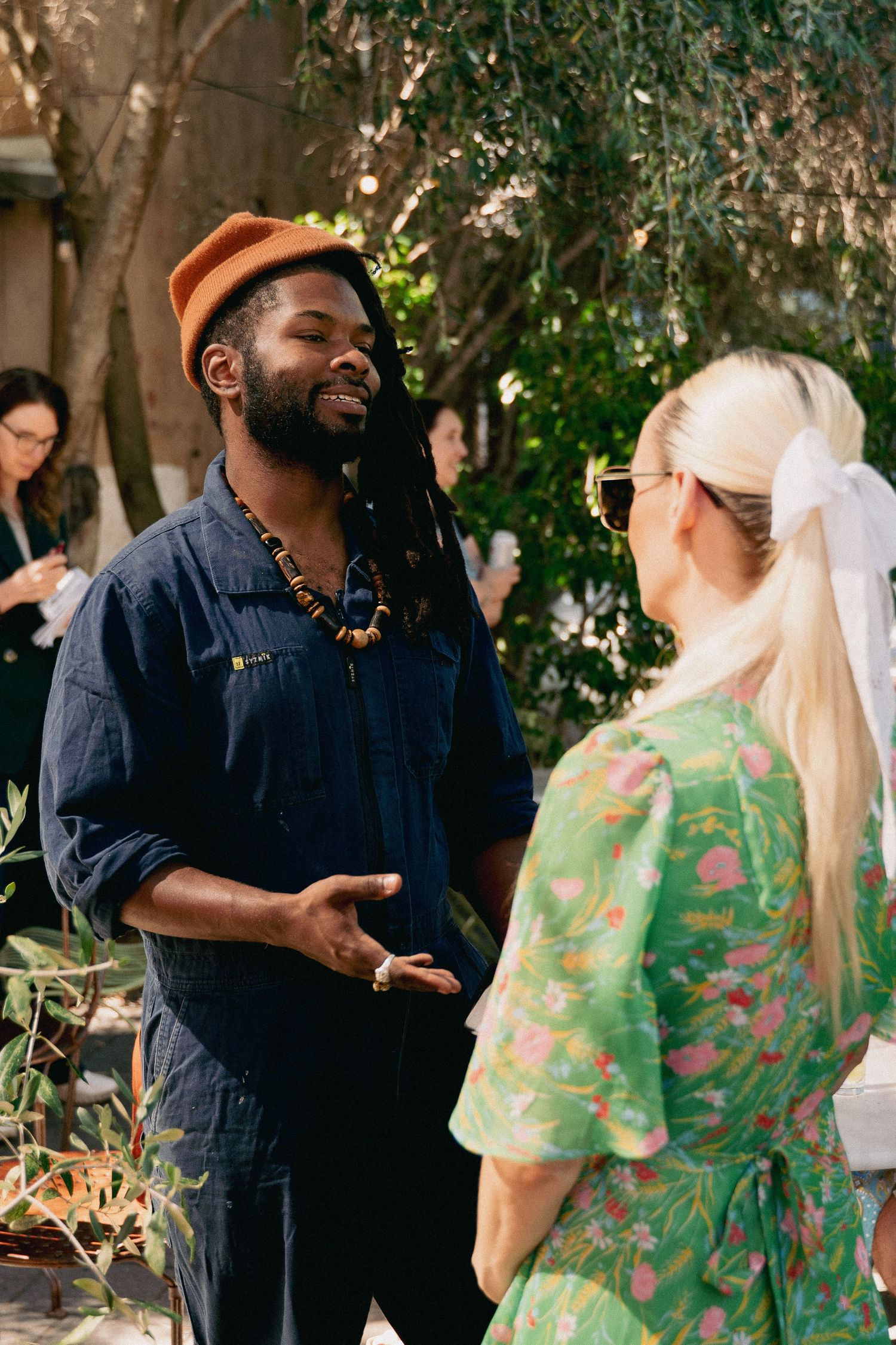 Man in orange hat with dreadlocks conversing with blonde woman in green floral dress.