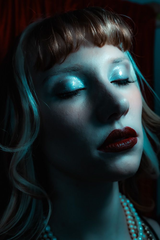Close-up portrait of a woman with teal eyeshadow, dark red lips, pearl necklace, and blonde hair in dramatic blue lighting.