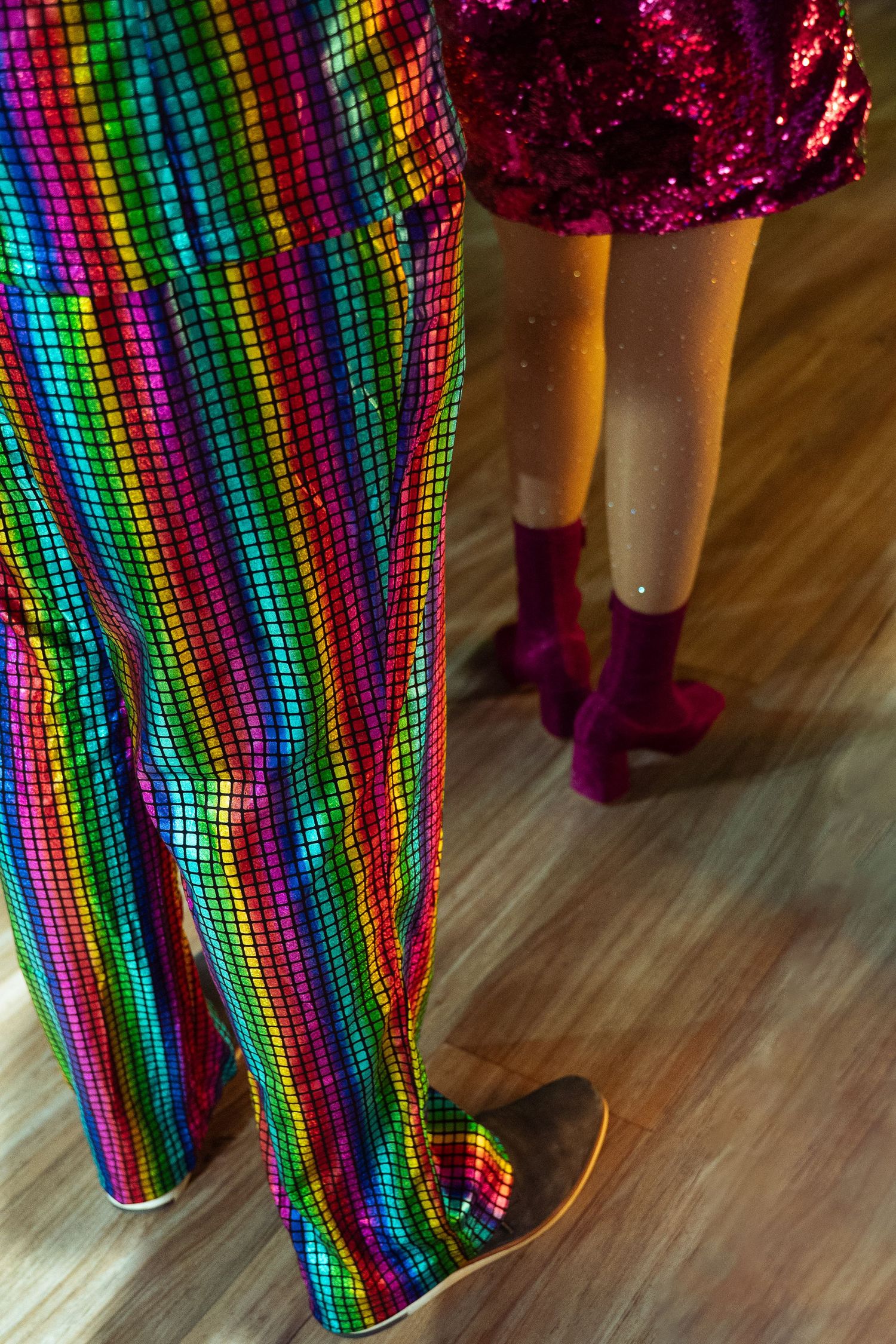 Two people wearing vibrant sequined outfits, rainbow mosaic pants and pink mini skirt, standing on wooden floor.
