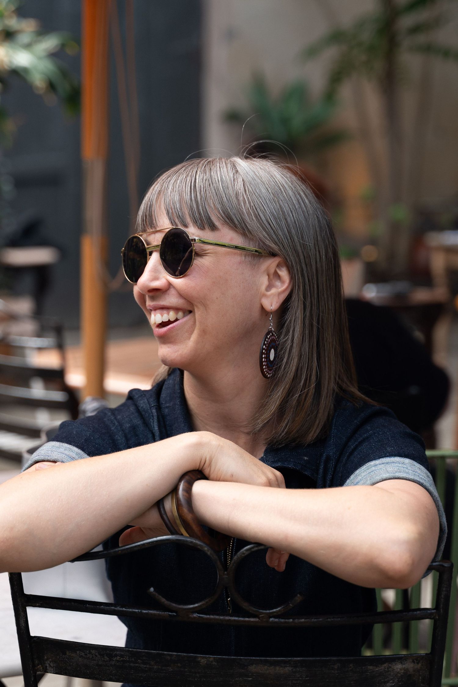 Smiling woman with bangs wearing sunglasses and earrings sits relaxed at an outdoor cafe chair.