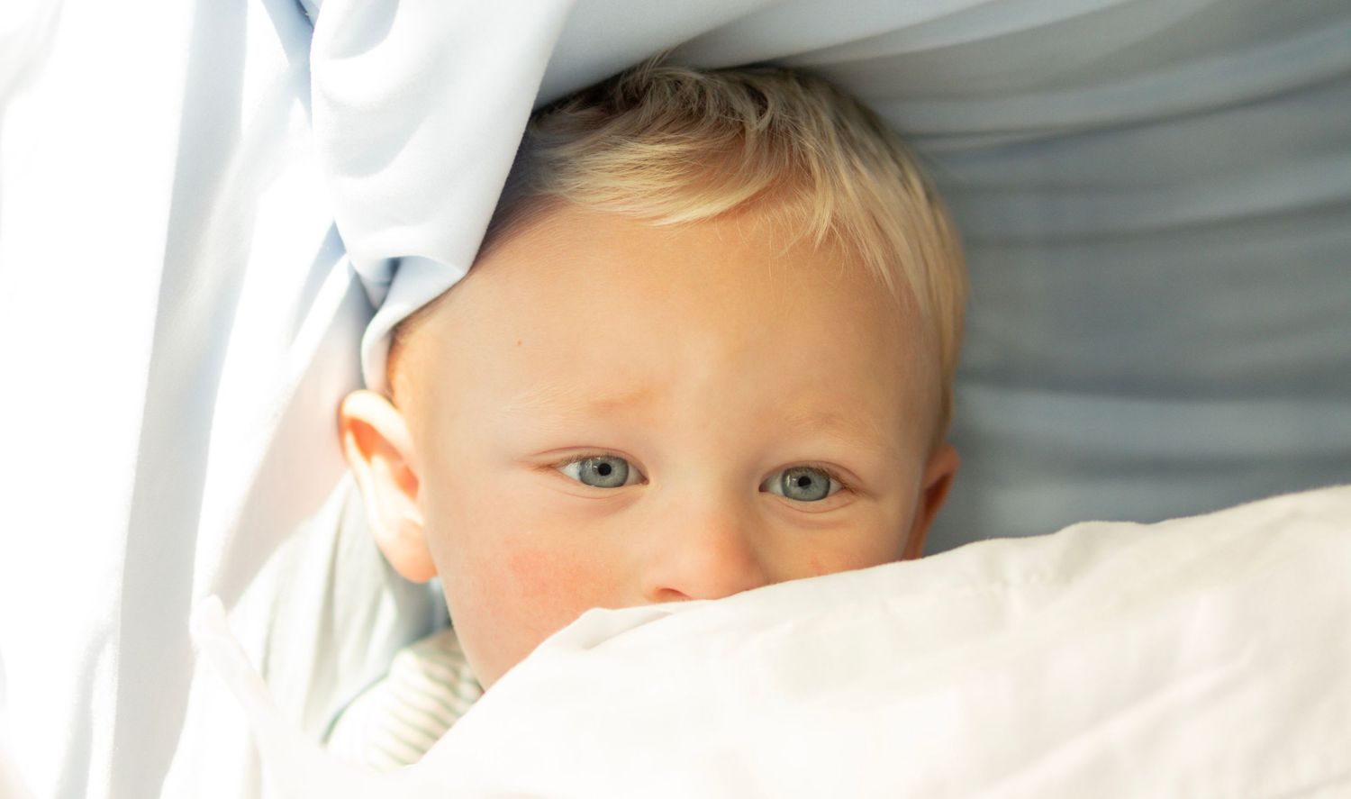 Adorable blue-eyed baby peeks curiously from beneath soft white and blue blankets.