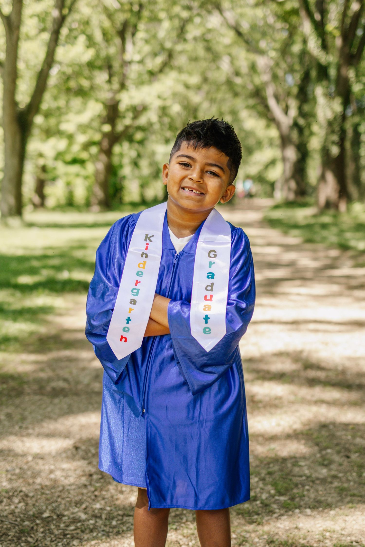 Happy boy in blue kindergarten graduation cap and gown with sash posing in tree-lined park path.