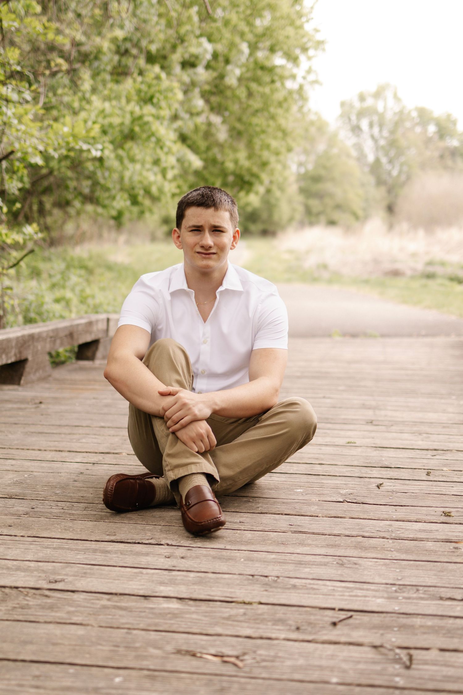 Young man in white shirt and khaki pants sits cross-legged on wooden bridge in nature park.