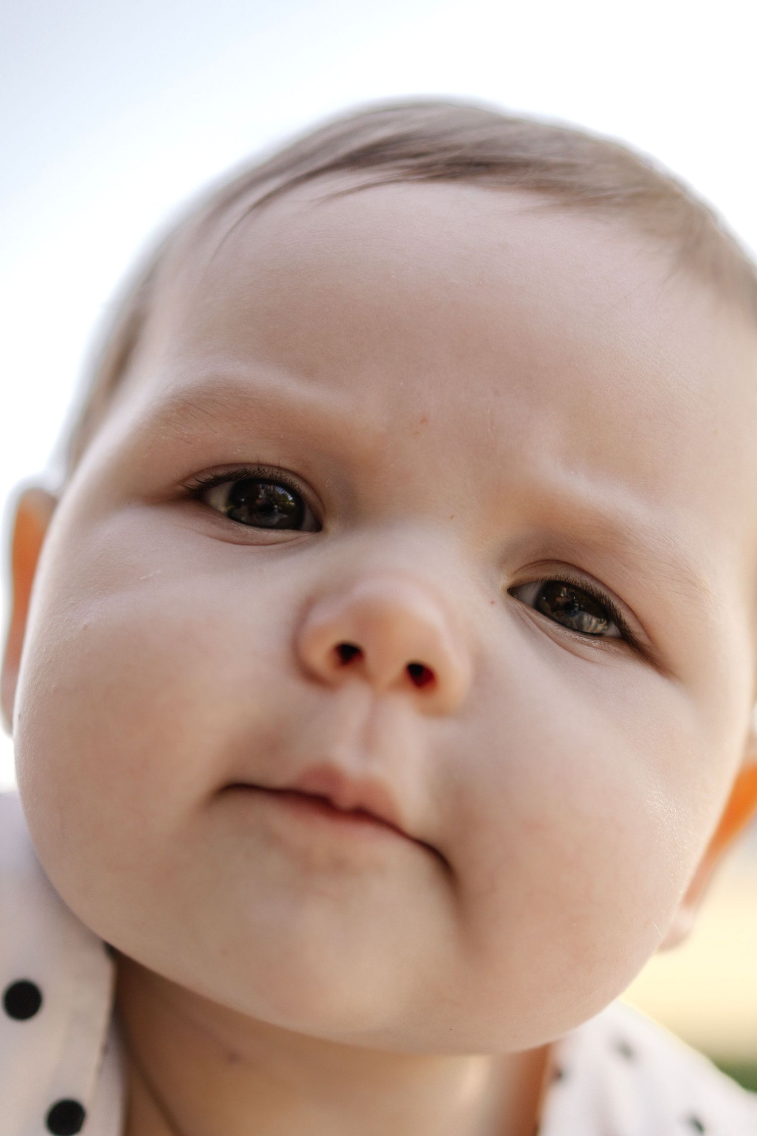 Extreme close-up portrait of chubby cheeked baby with dark eyes looking up against bright sky.