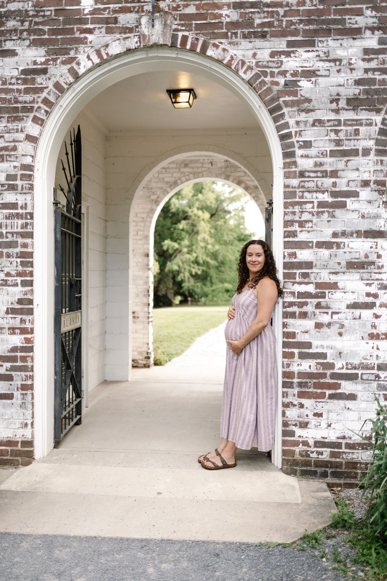 Smiling pregnant woman in striped maxi dress stands in brick archway holding baby bump.