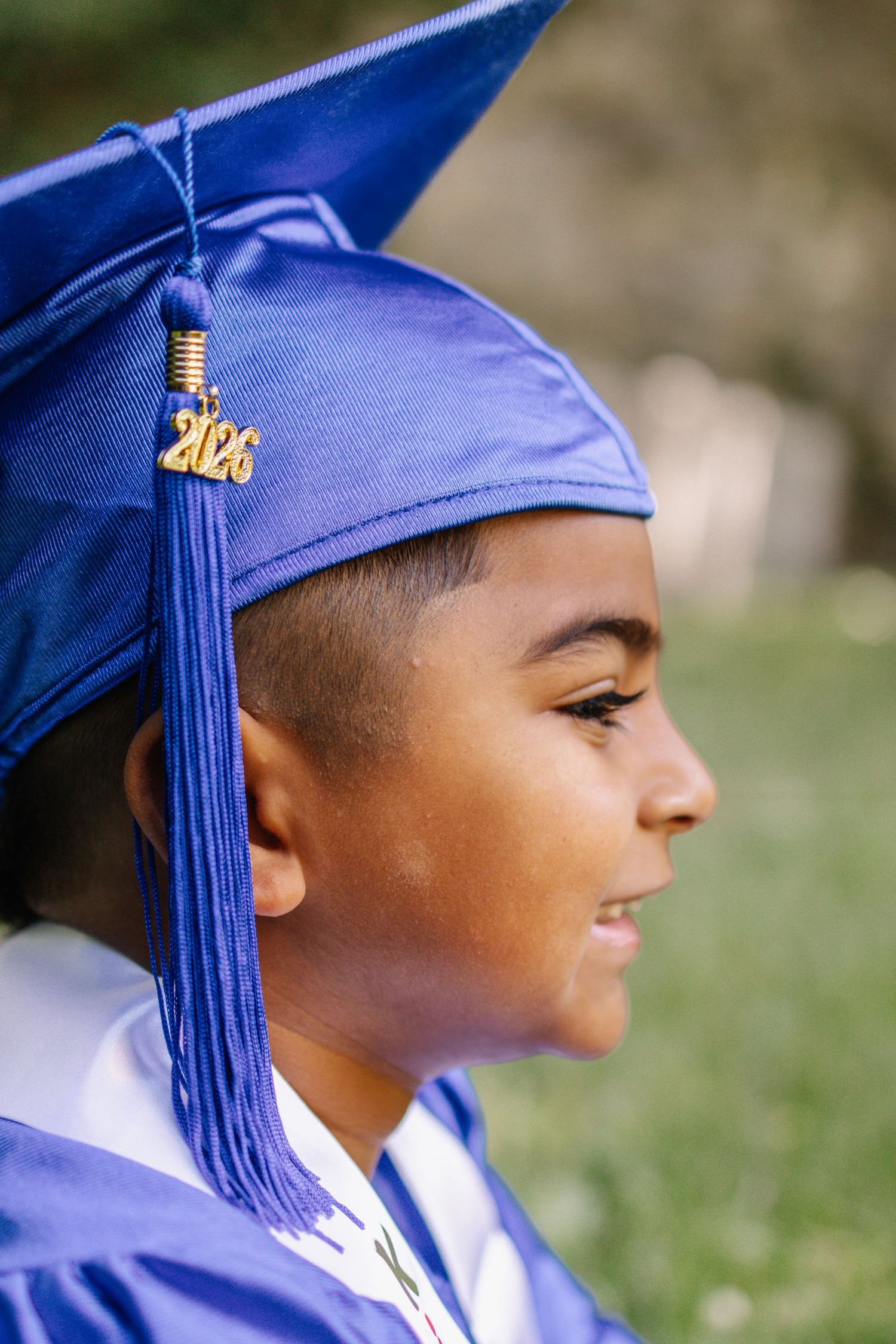 Side profile of young boy wearing blue graduation cap with 2026 tassel smiling outdoors.