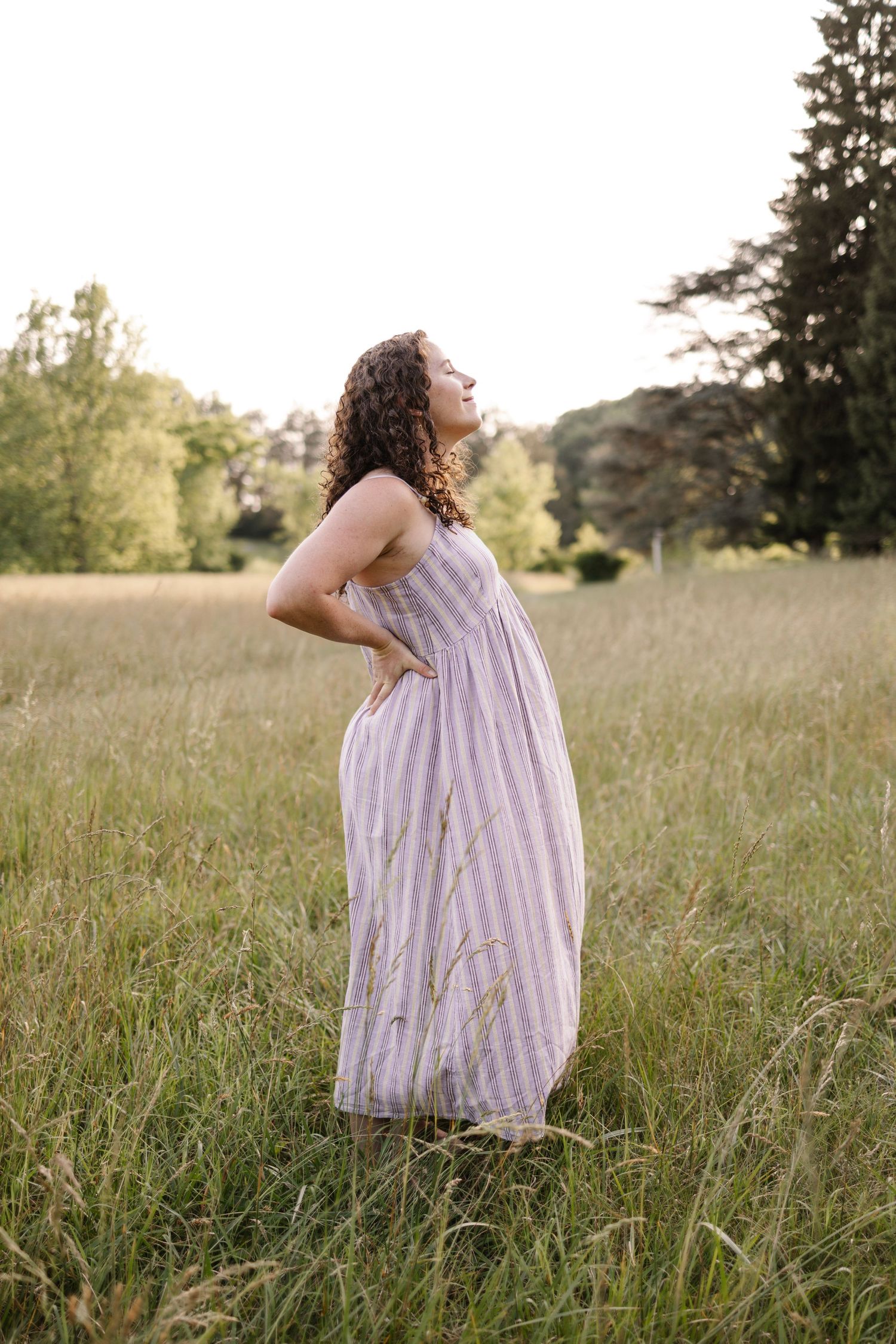 Pregnant woman in striped maxi dress stands in golden meadow with hands on back at sunset.
