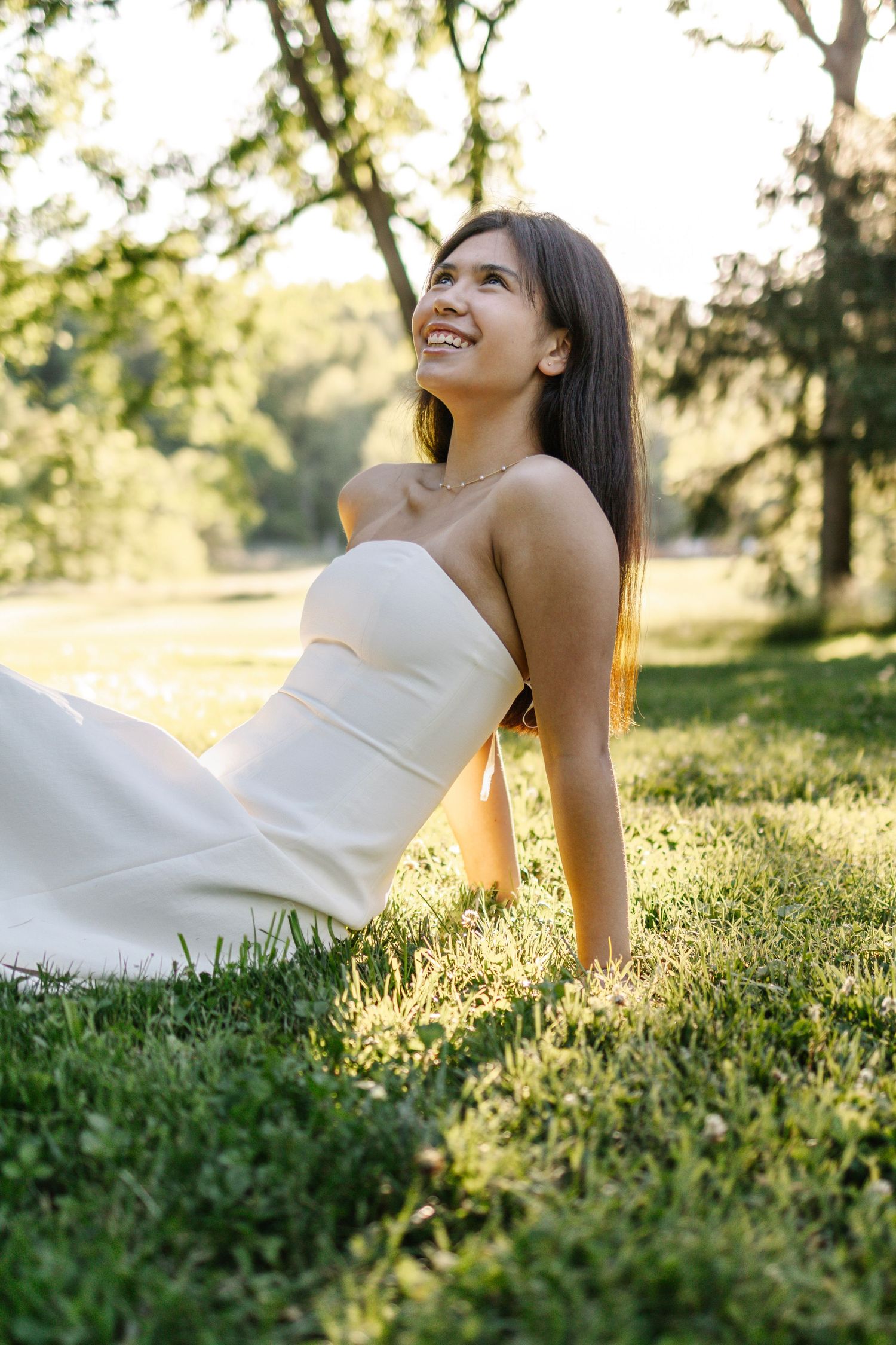 Beautiful brunette bride in strapless white wedding gown sitting on grass smiling in sunlit park.