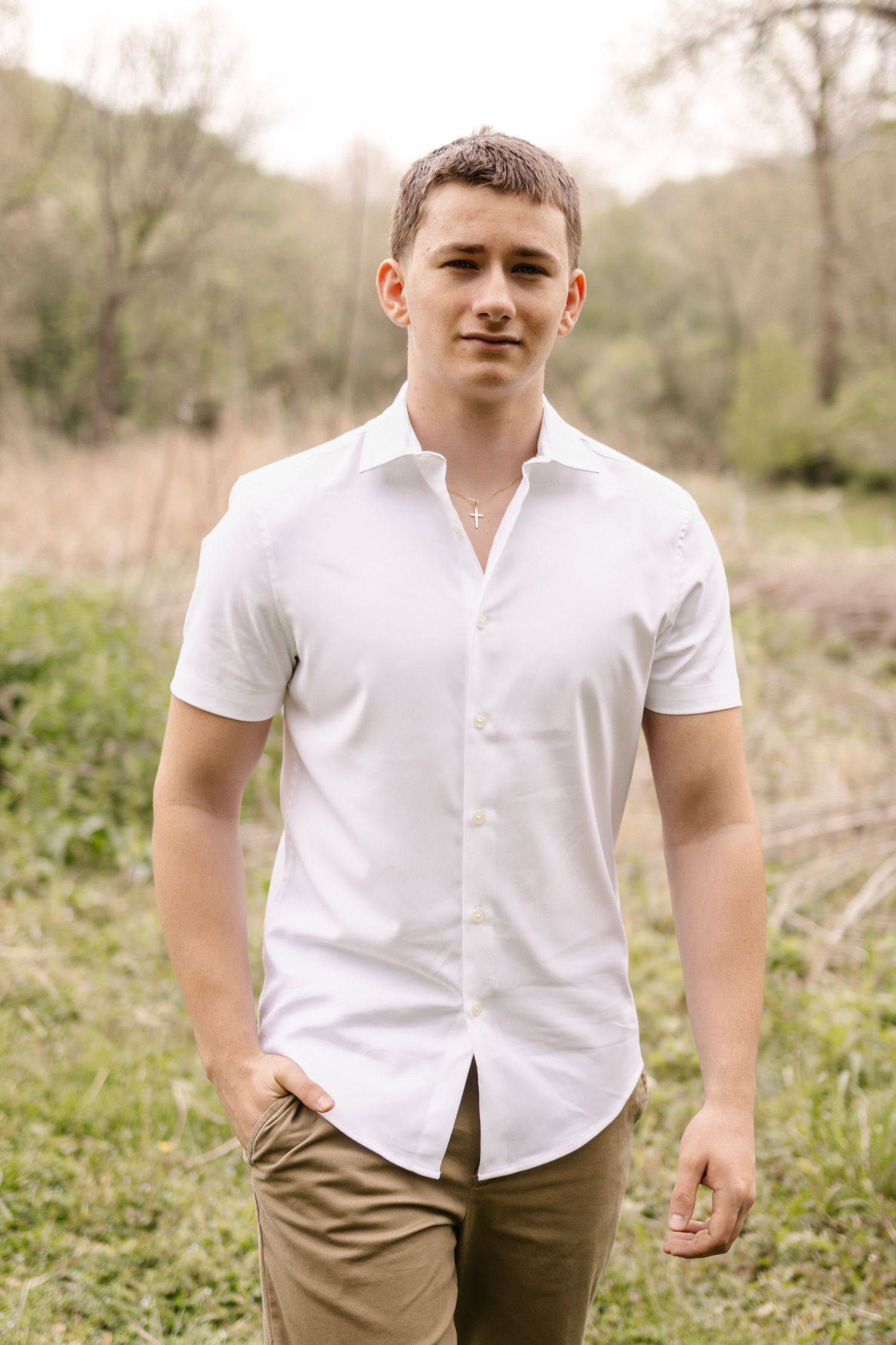 Young man in white short-sleeve button-up shirt and khaki pants stands in natural outdoor setting.