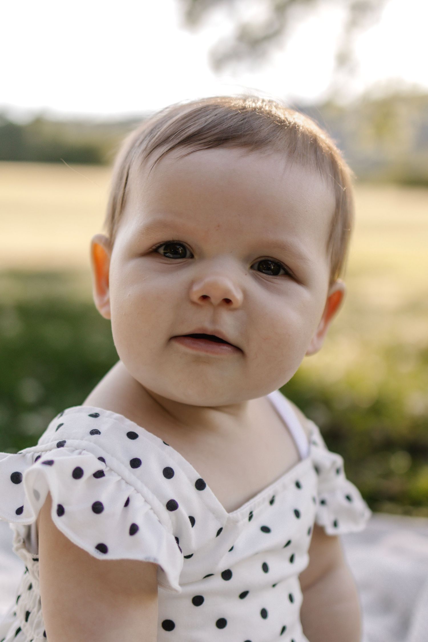 Close-up portrait of cute baby girl in white polka dot dress outdoors in sunlit park setting.