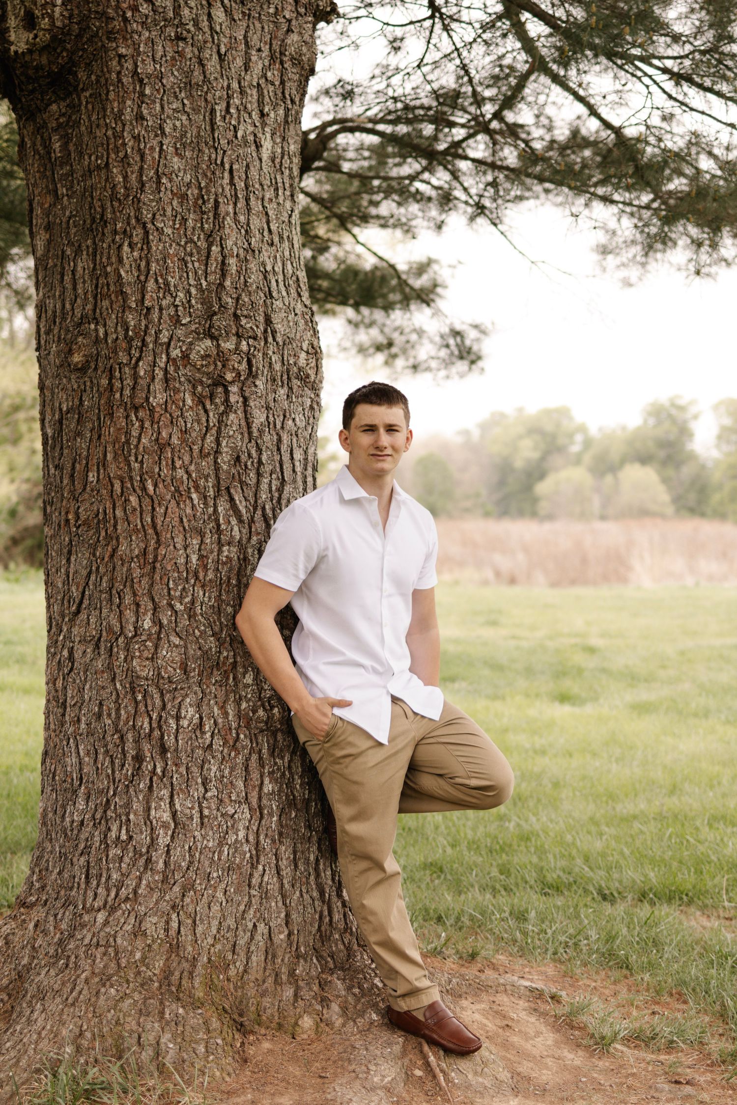 Young man in white shirt and khaki pants leans casually against large tree trunk in green field.