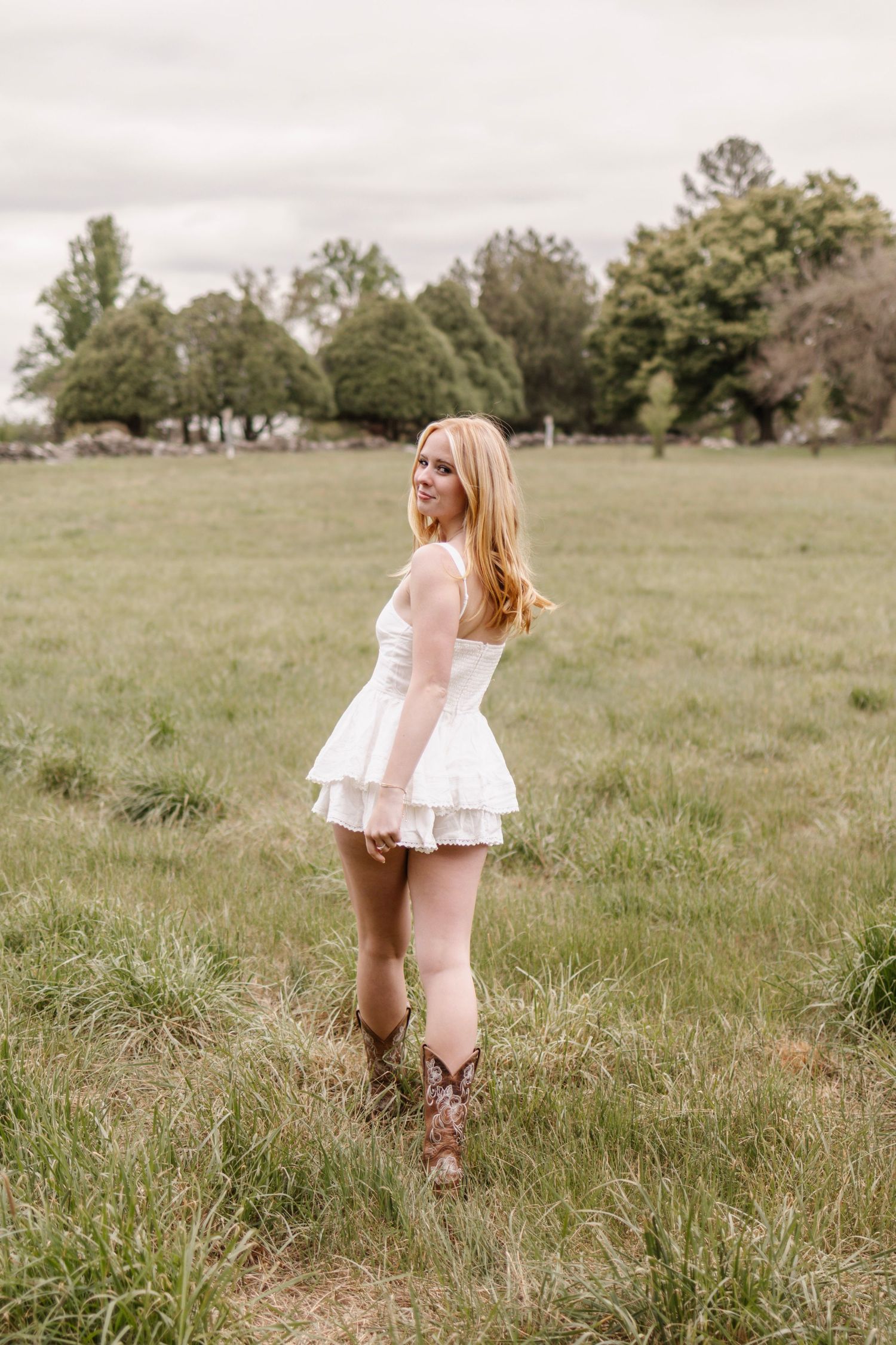 Blonde woman in white mini dress and cowboy boots standing in a green field on a cloudy day.