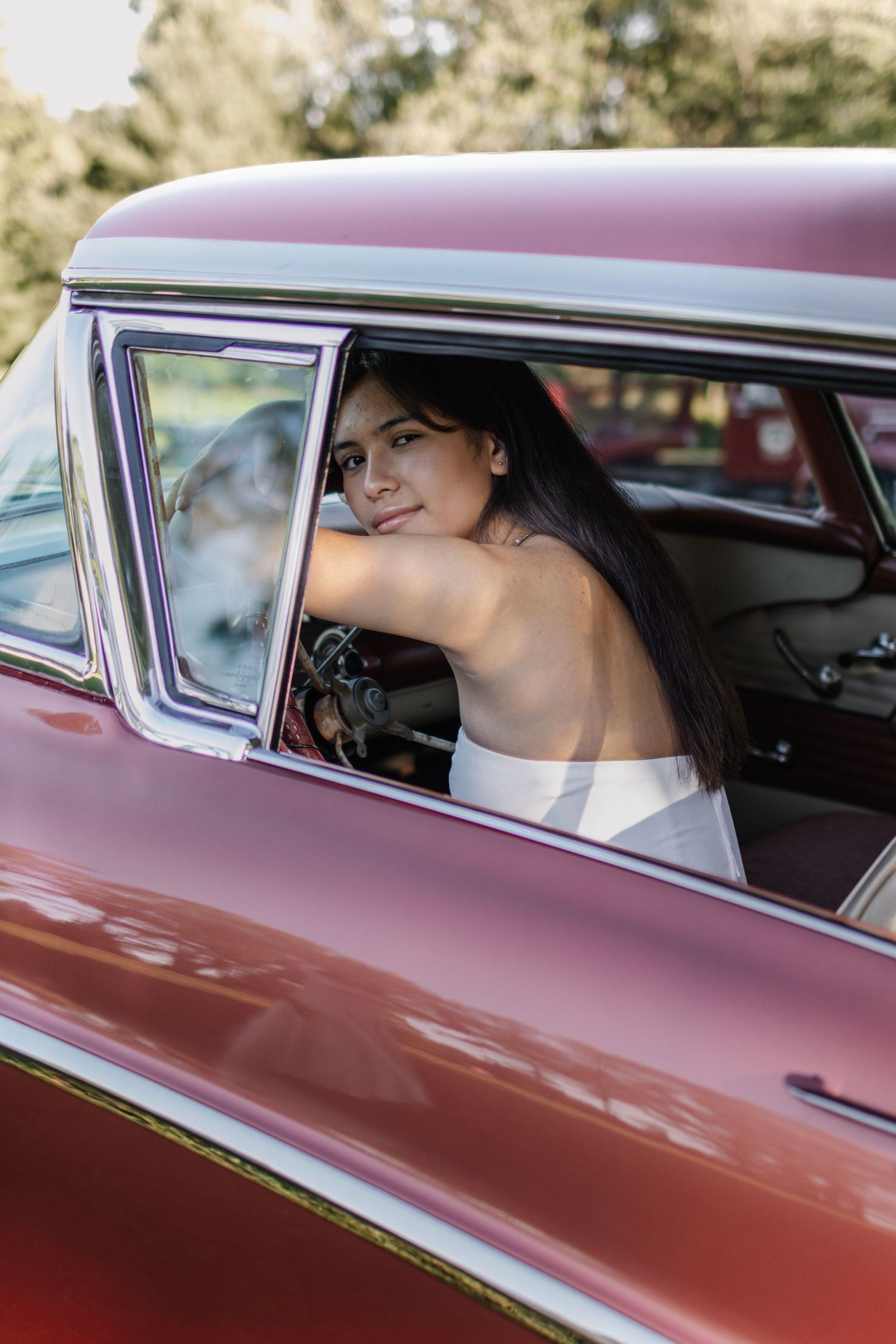 Smiling brunette woman in white dress leans out window of vintage red classic car outdoors.