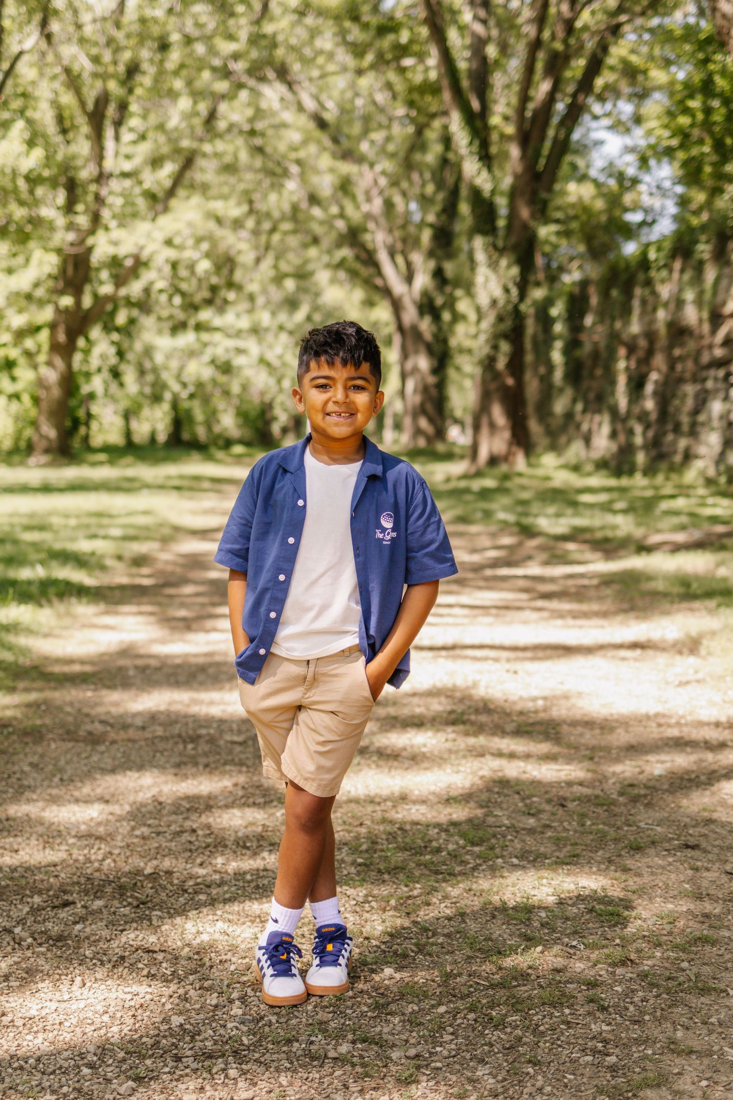 Smiling young boy in blue shirt and khaki shorts standing on shaded park path with hands in pockets.