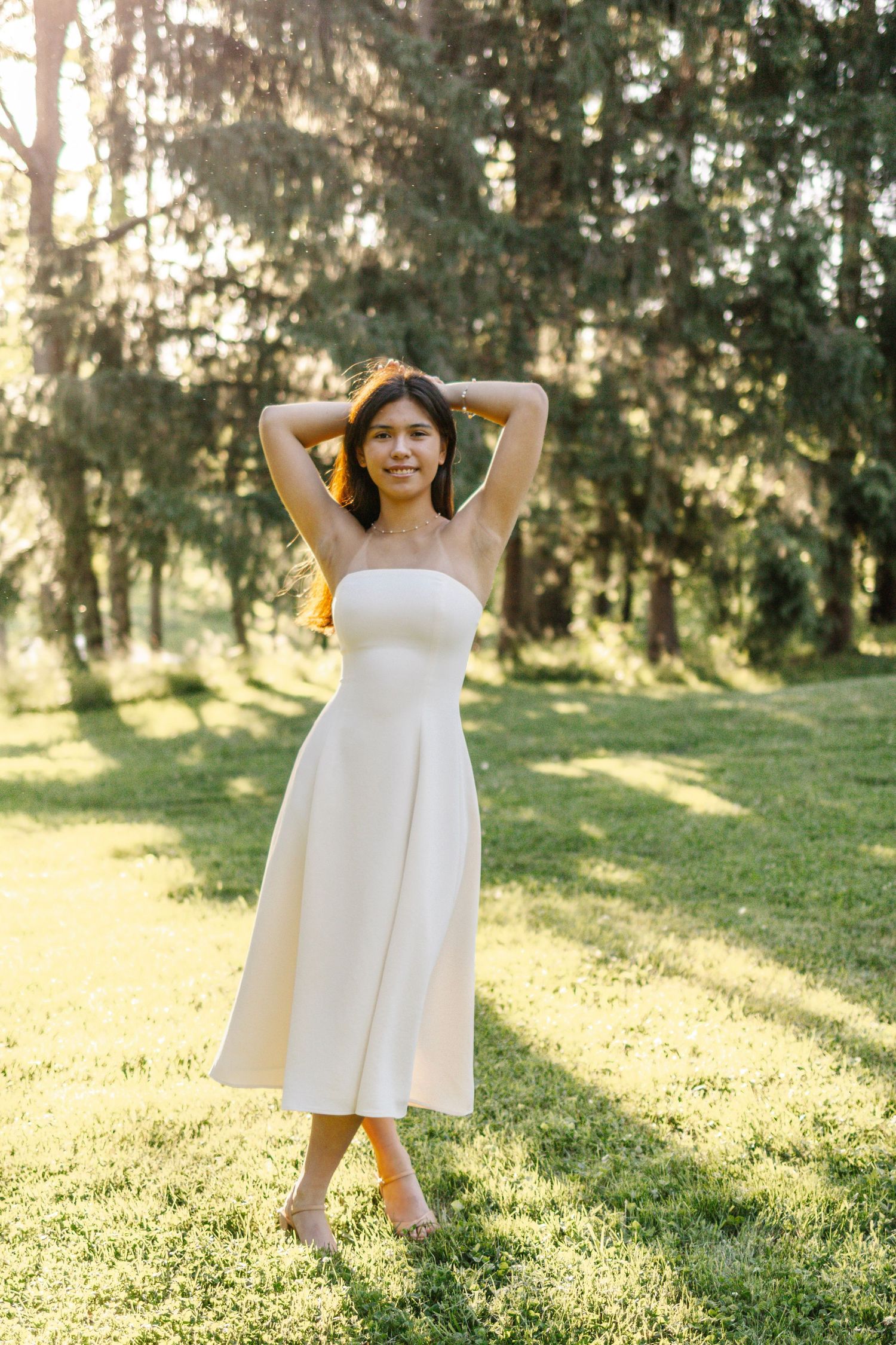 Young woman in white strapless midi dress poses gracefully in sunlit park with hands behind head.