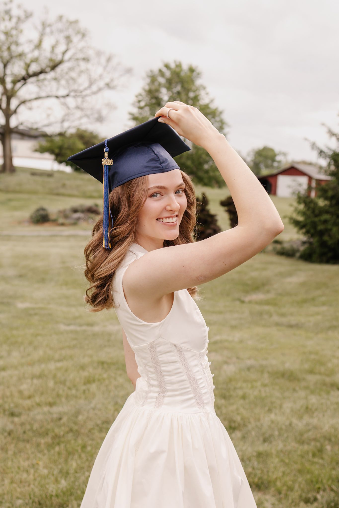Joyful female graduate in white dress adjusting navy blue graduation cap outdoors on green lawn.
