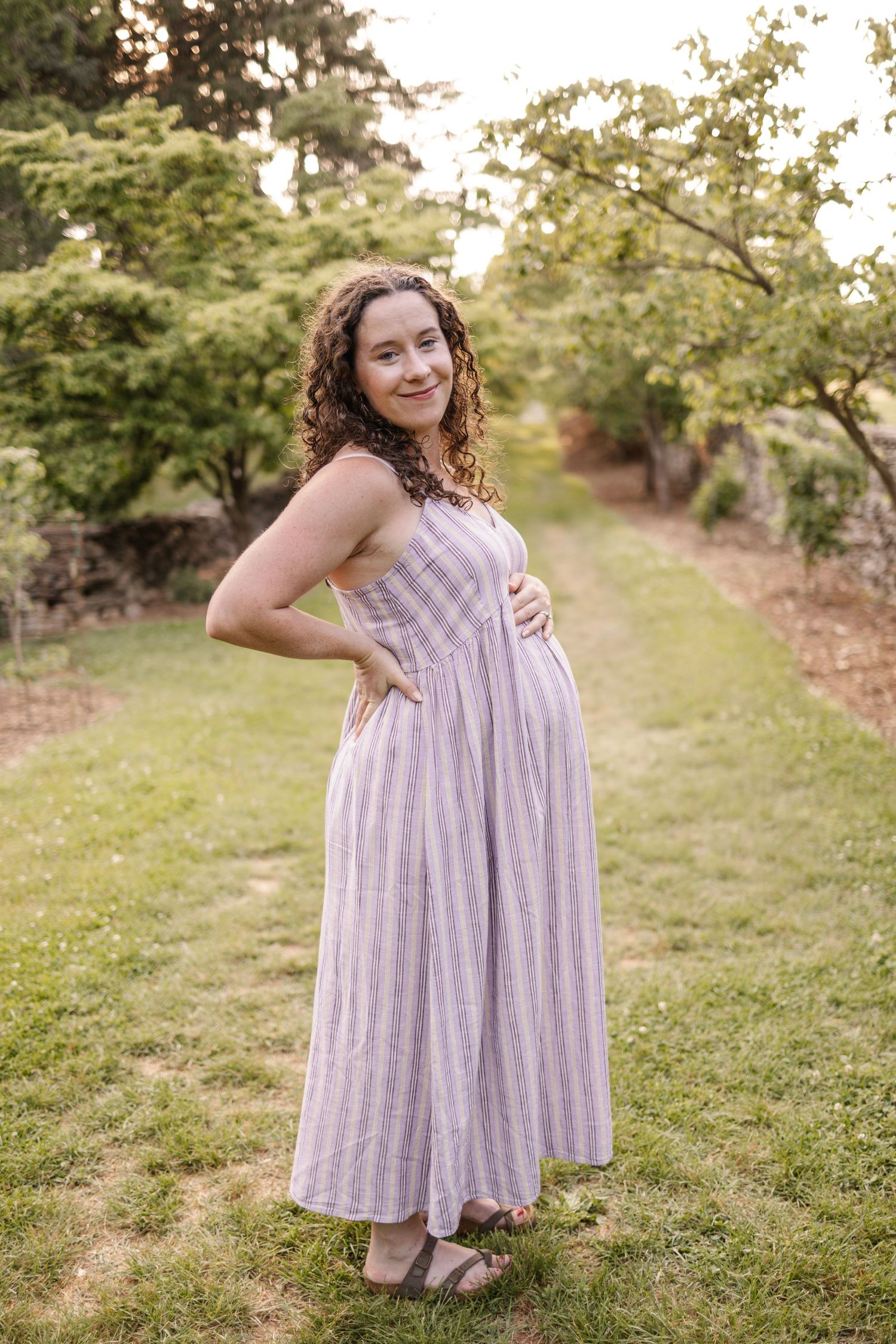 Smiling pregnant woman in striped lavender maxi dress poses in lush green garden pathway.