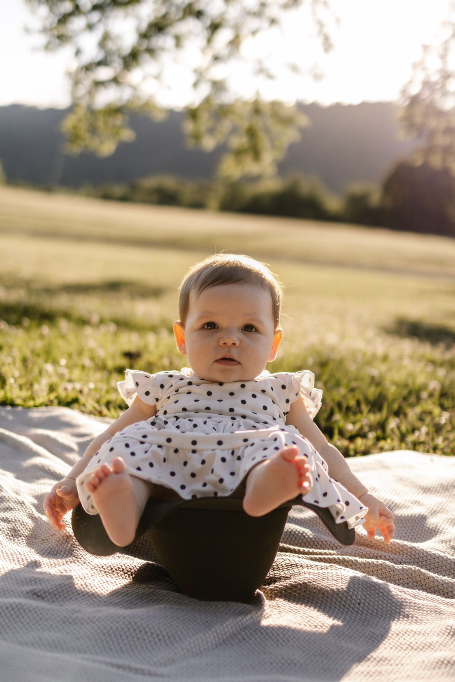 Adorable baby girl in polka dot dress sitting on blanket in sunny green field during golden hour.