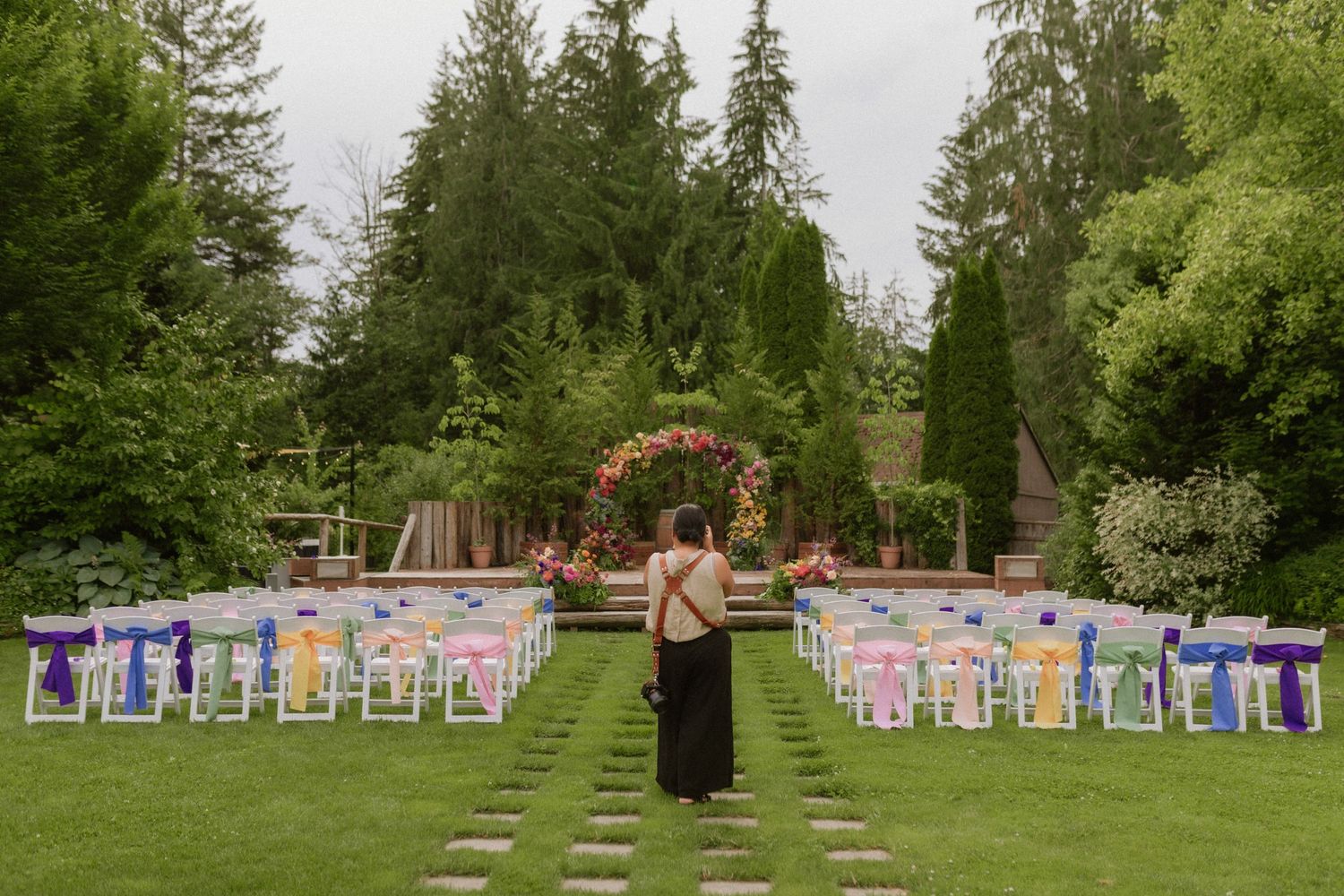 Outdoor wedding setup with rows of colorful chairs leading to a floral arch in a green garden setting.