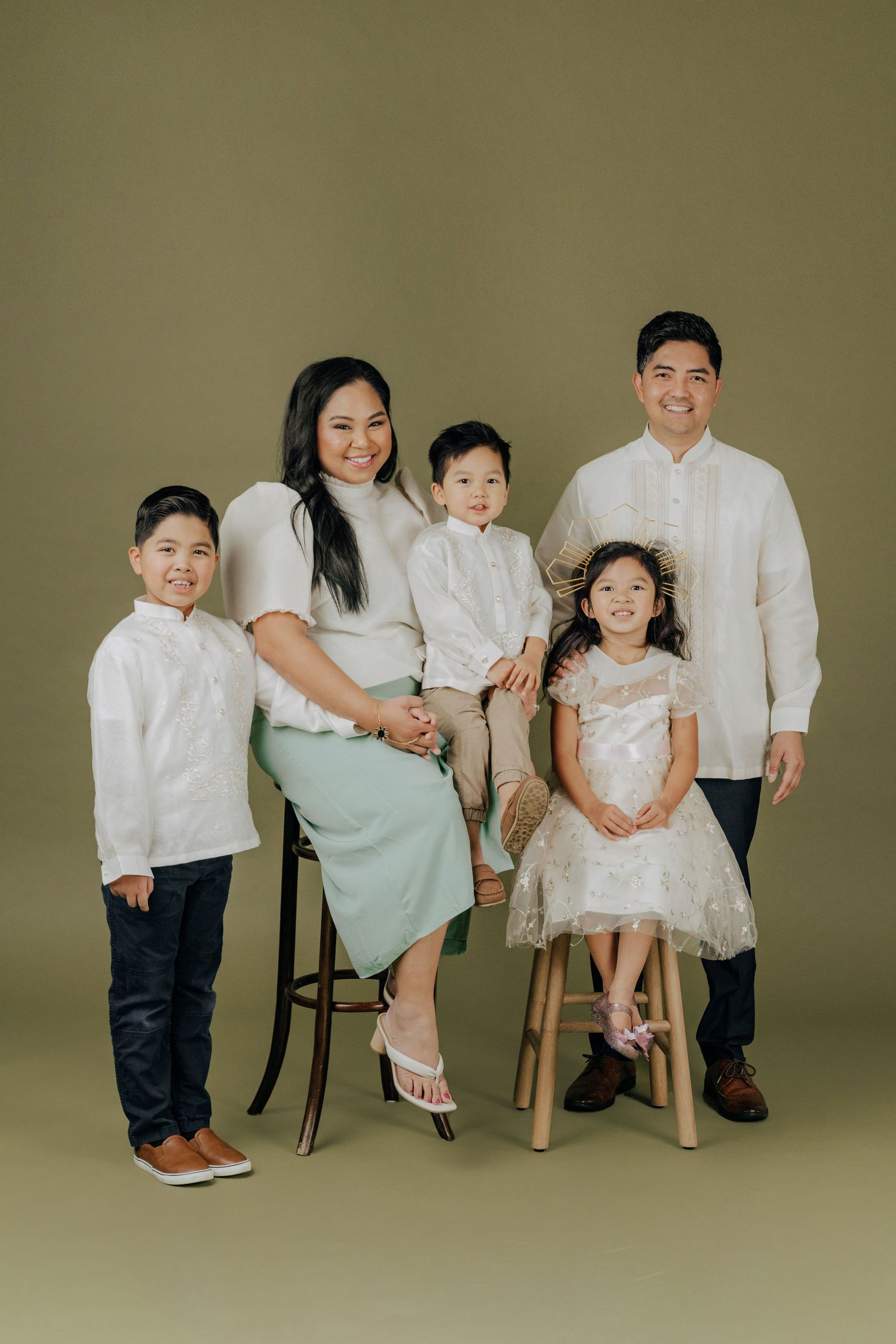 Studio family portrait with coordinated white and neutral outfits against beige backdrop.